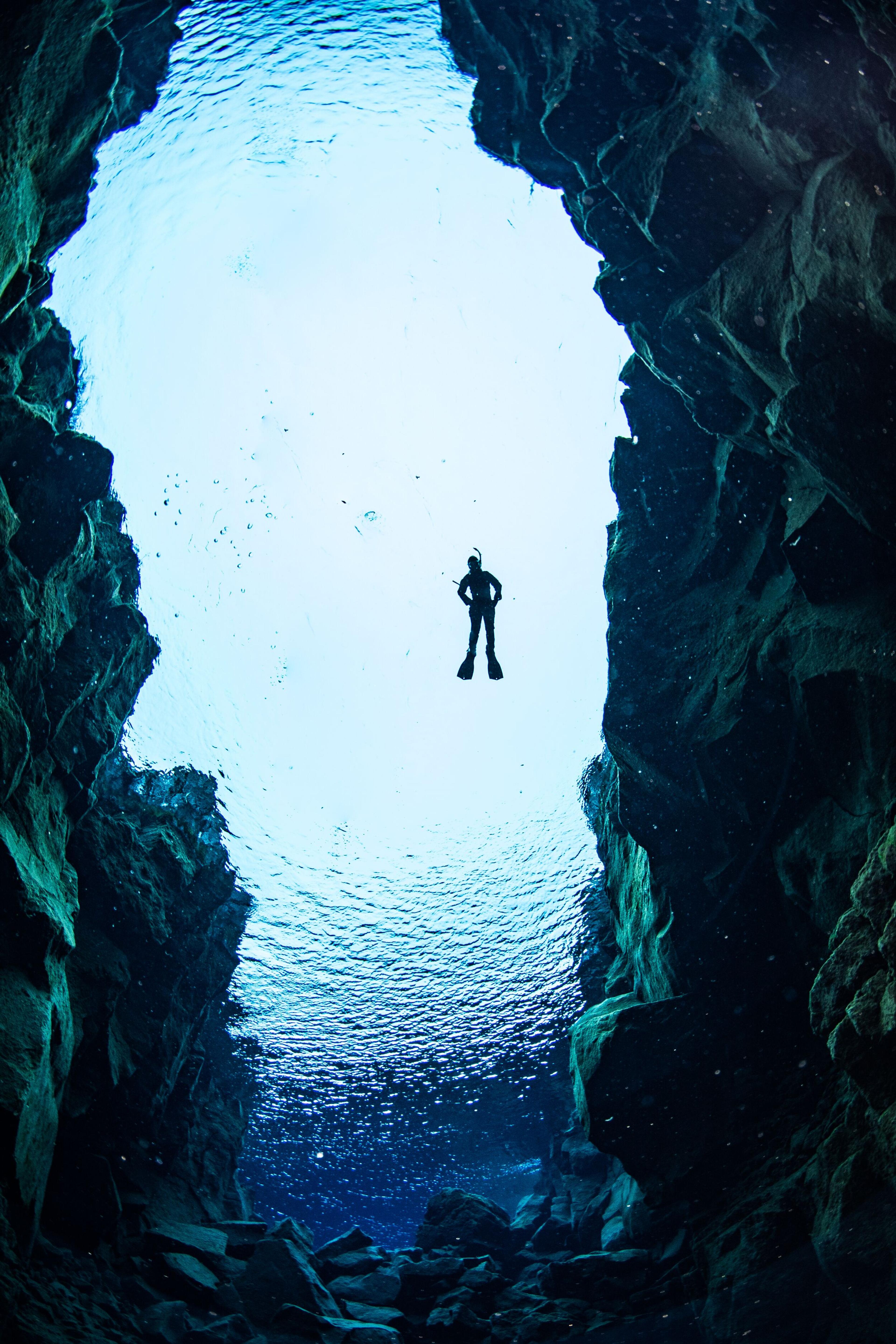 Silhouetted diver framed by dark rock walls, seen from below in clear blue water.