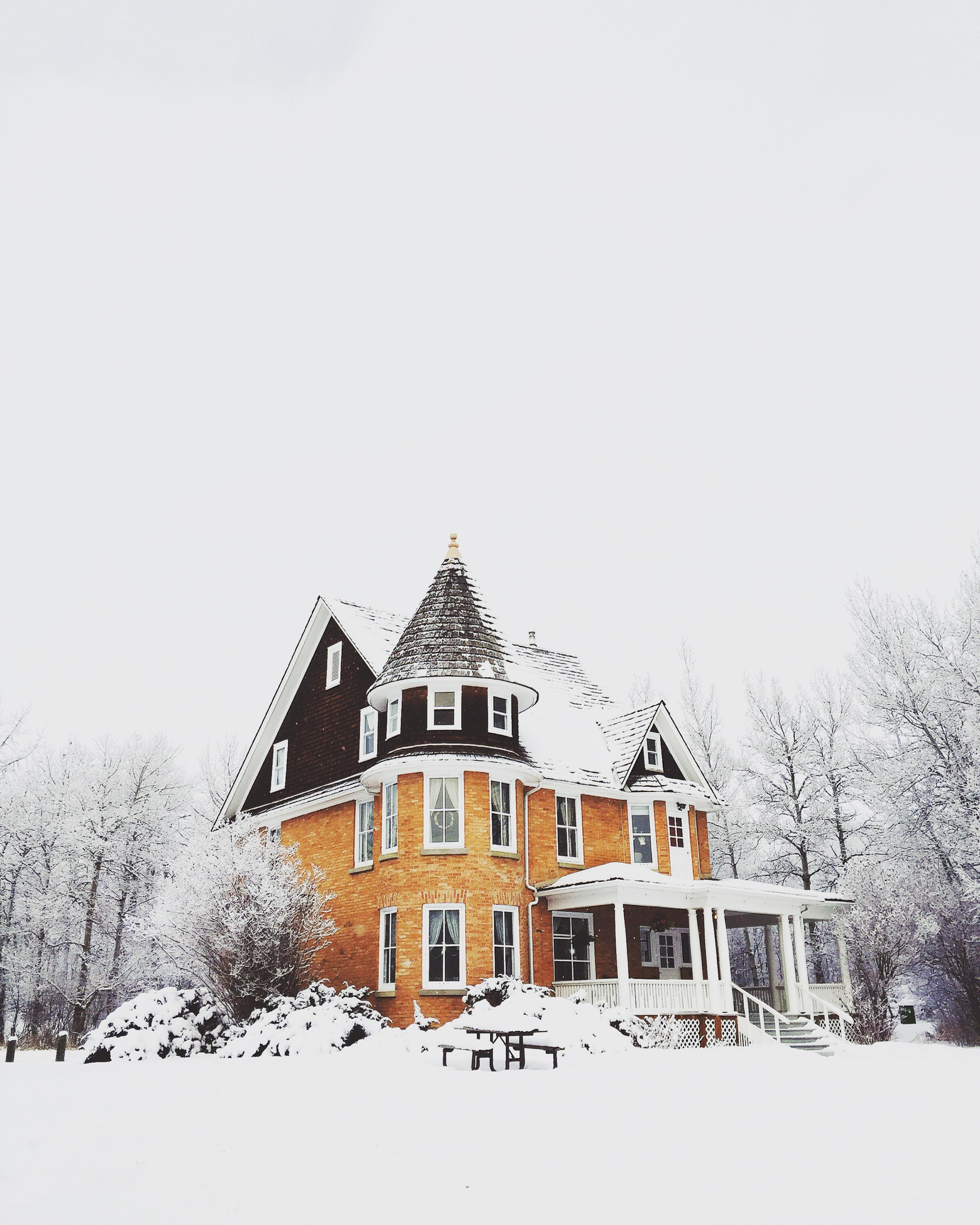 A two-story orange house with a pointed roof, surrounded by snow-covered trees and a white snowy ground, under a cloudy winter sky.