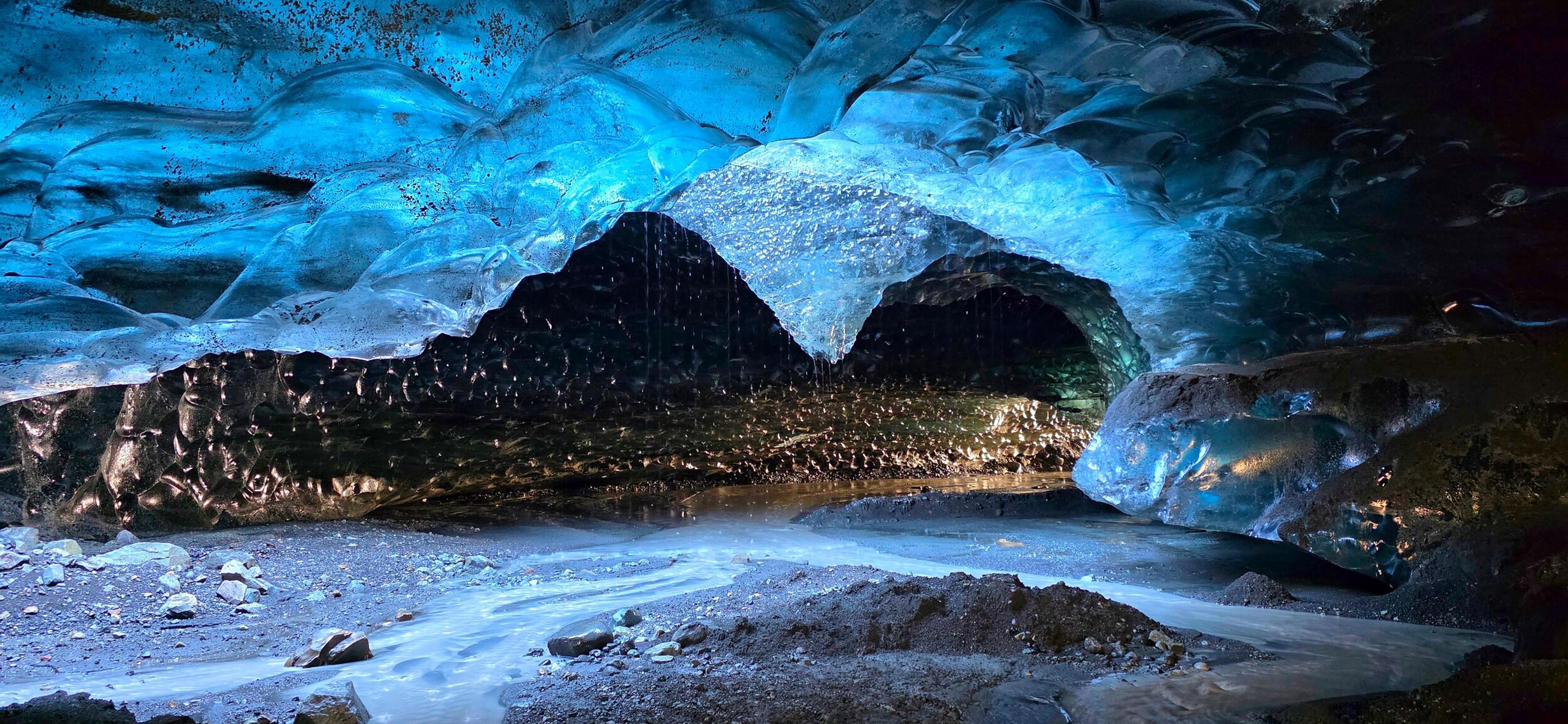 Vibrant blue ice cave interior with a stream flowing over a dark, rocky floor.