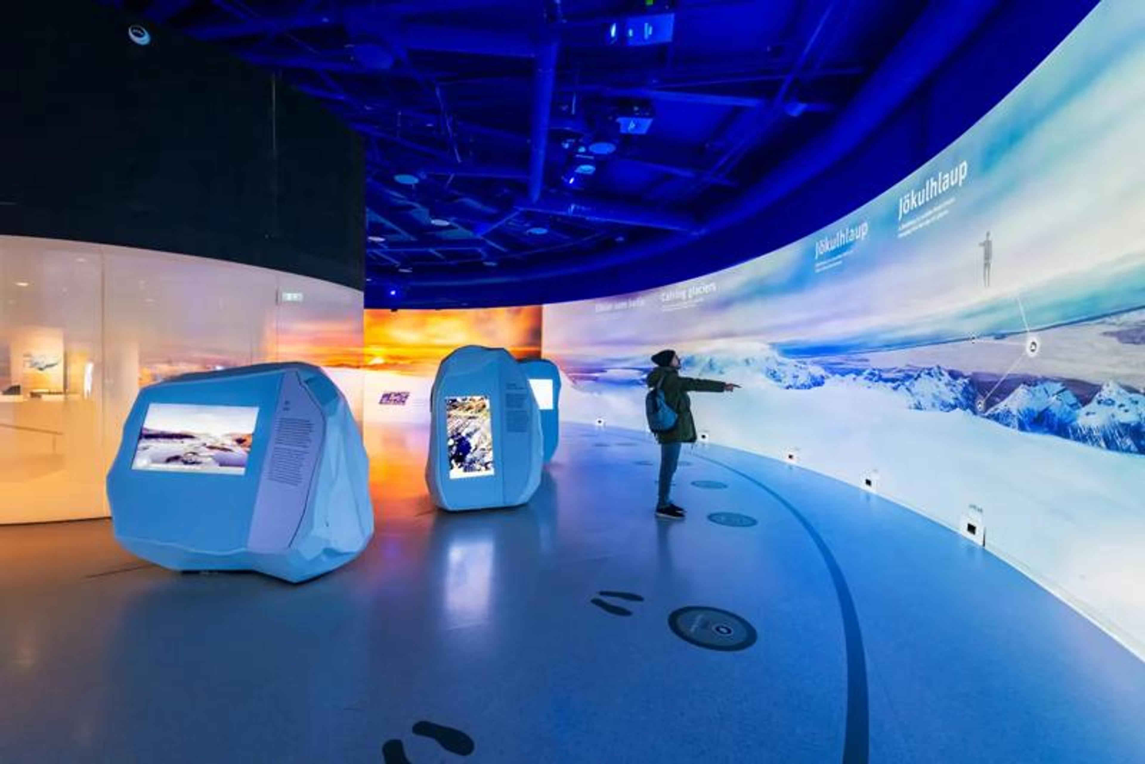 A person points at a large curved screen displaying a glacier landscape in a blue-lit museum exhibit with rock-shaped interactive displays.