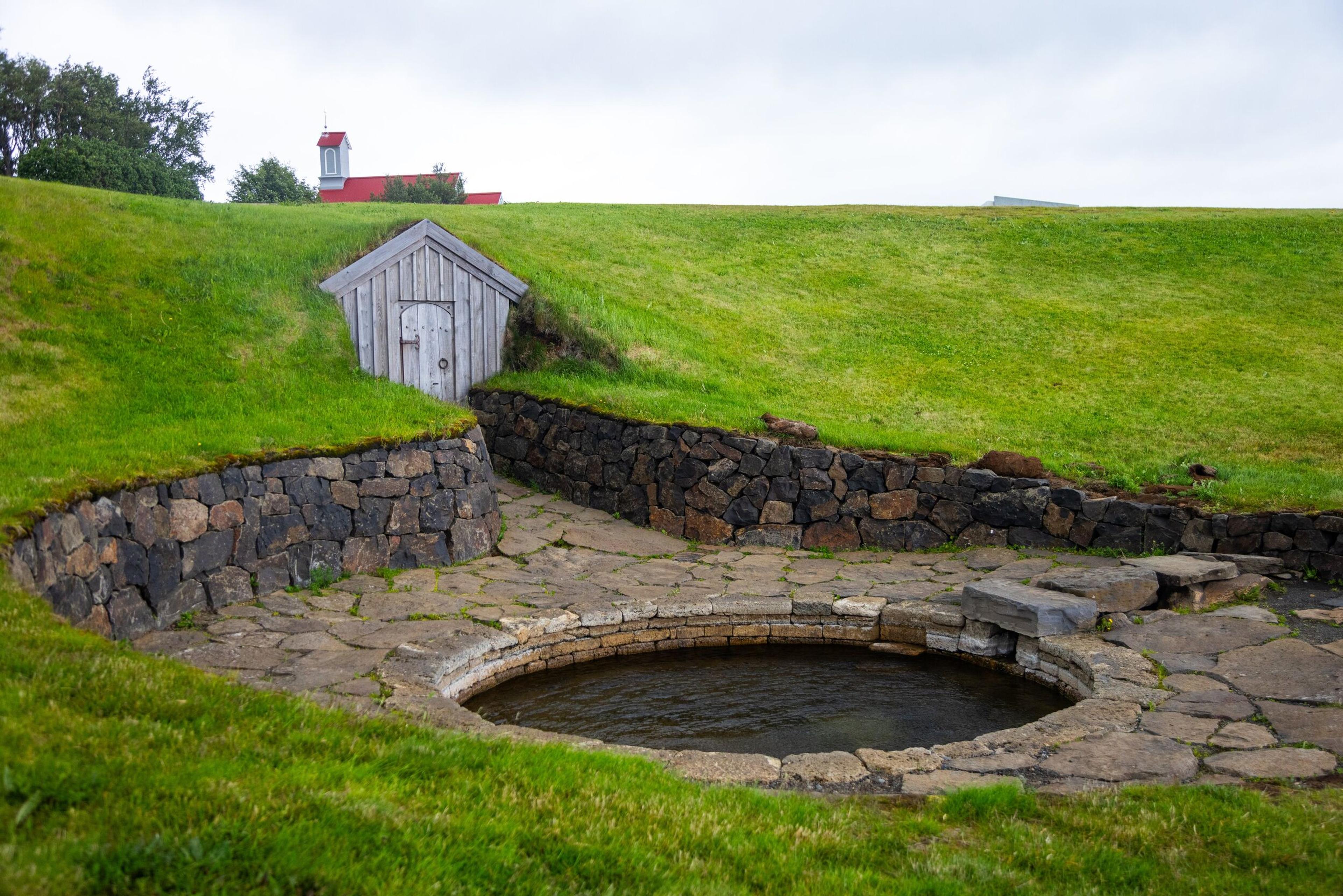 Snorralaug, a historic circular stone hot spring in Reykholt, surrounded by green grass and a small wooden shed, reflecting Iceland's cultural heritage.