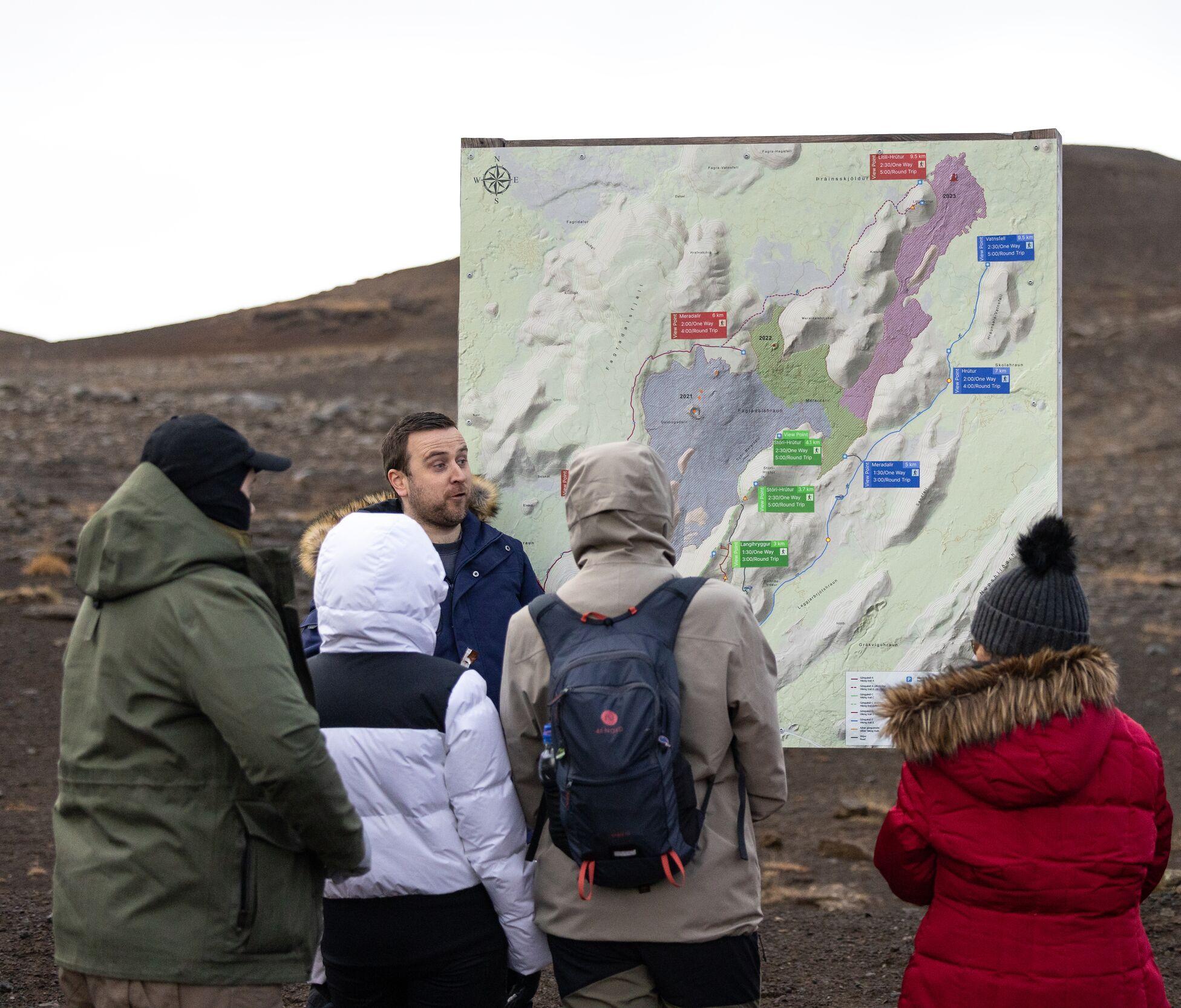 A group of people gathered around an informational map on the Reykjanes Peninsula, as a guide explains the layout and features of the volcanic area near Grindavik, with barren terrain in the background.