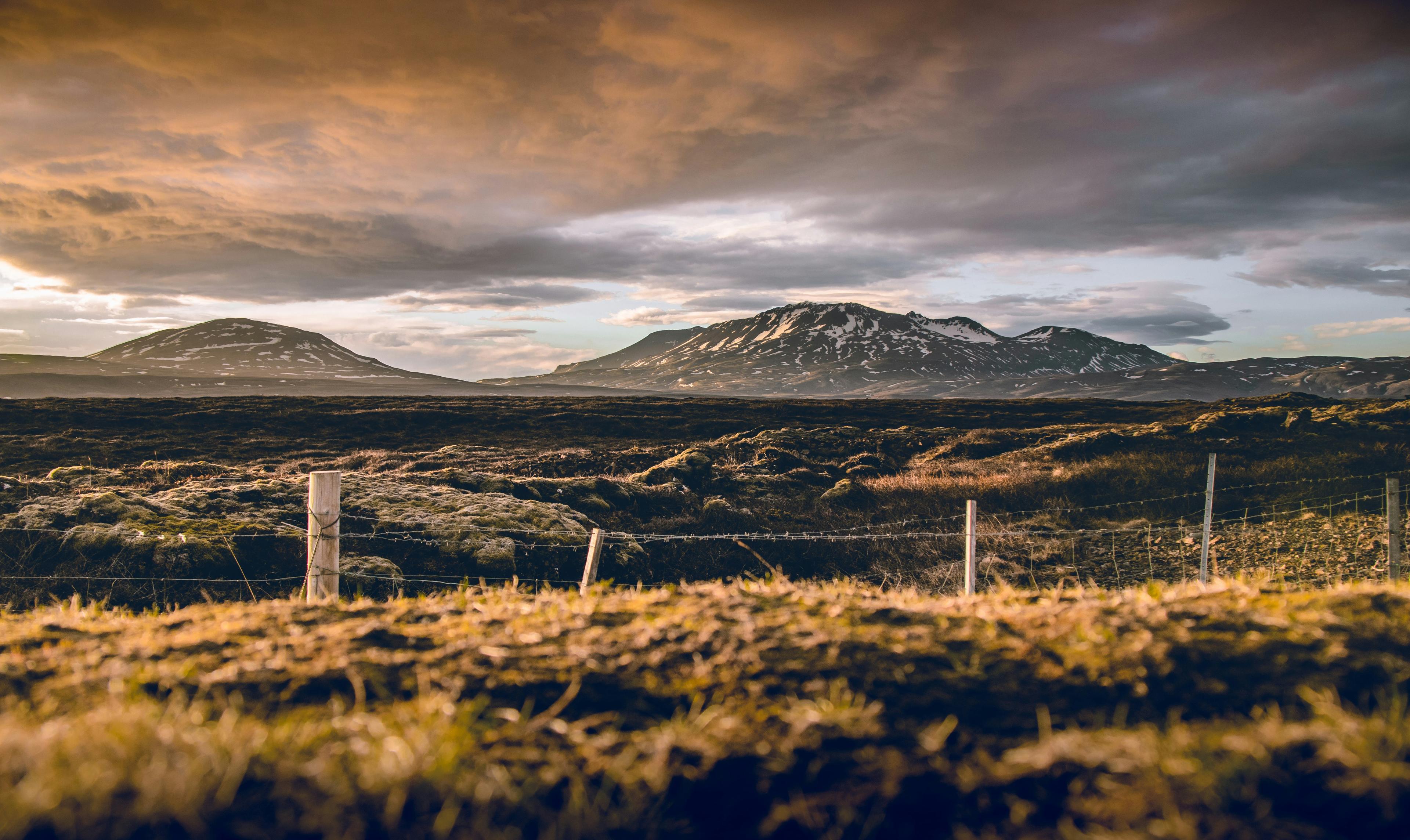 A moody sky looms over a rugged landscape with a fence in the foreground, leading the eye towards distant mountains bathed in the warm, golden light of a setting or rising sun.