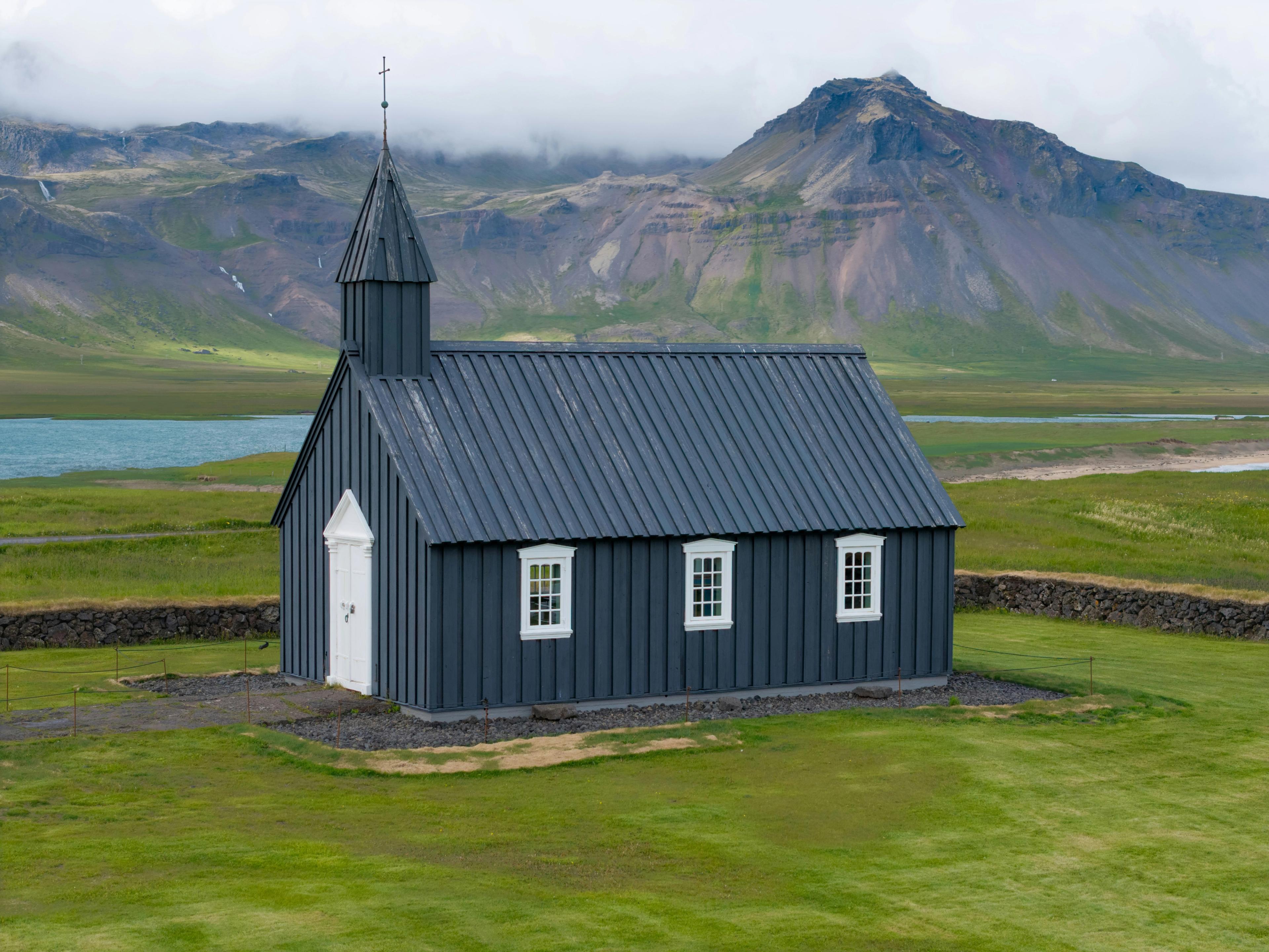 Black church with white trim in a green field, backed by a lake and cloudy mountains.