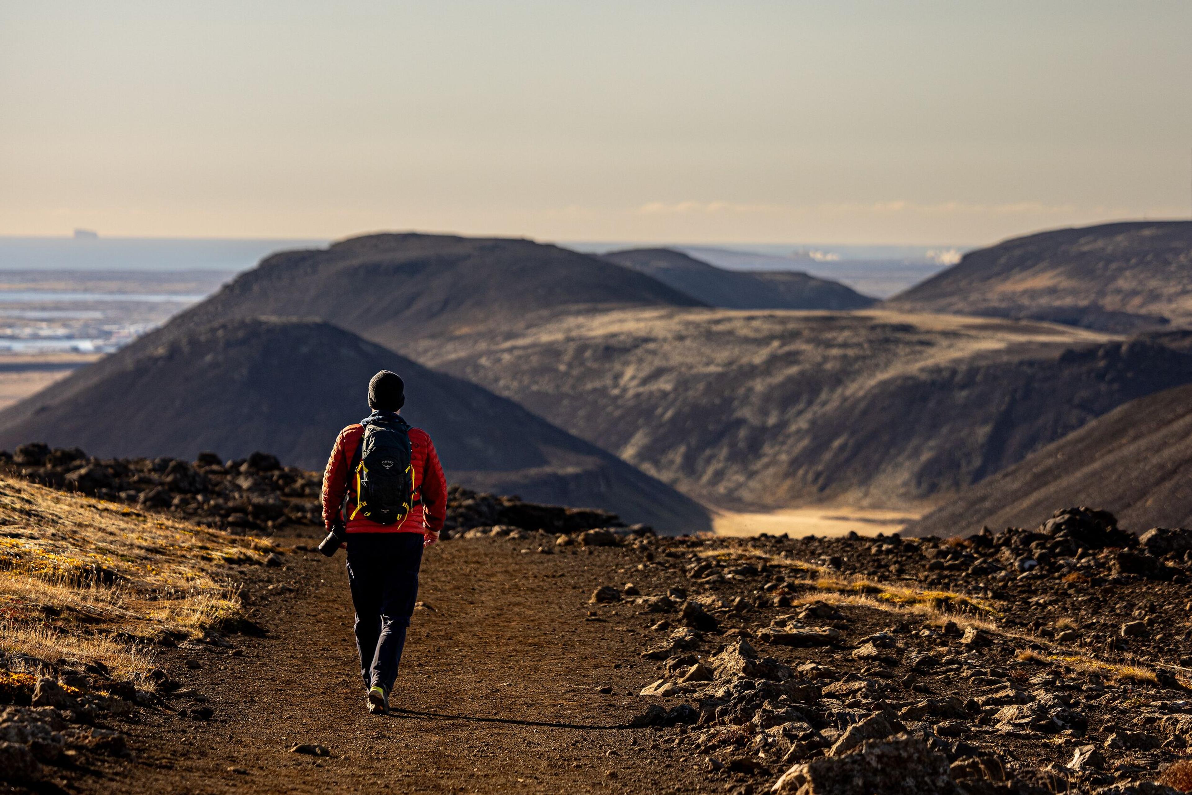 A hiker in a red jacket and backpack explores a rocky trail through the Reykjanes volcanic terrain, with scenic hills and valleys in the background.