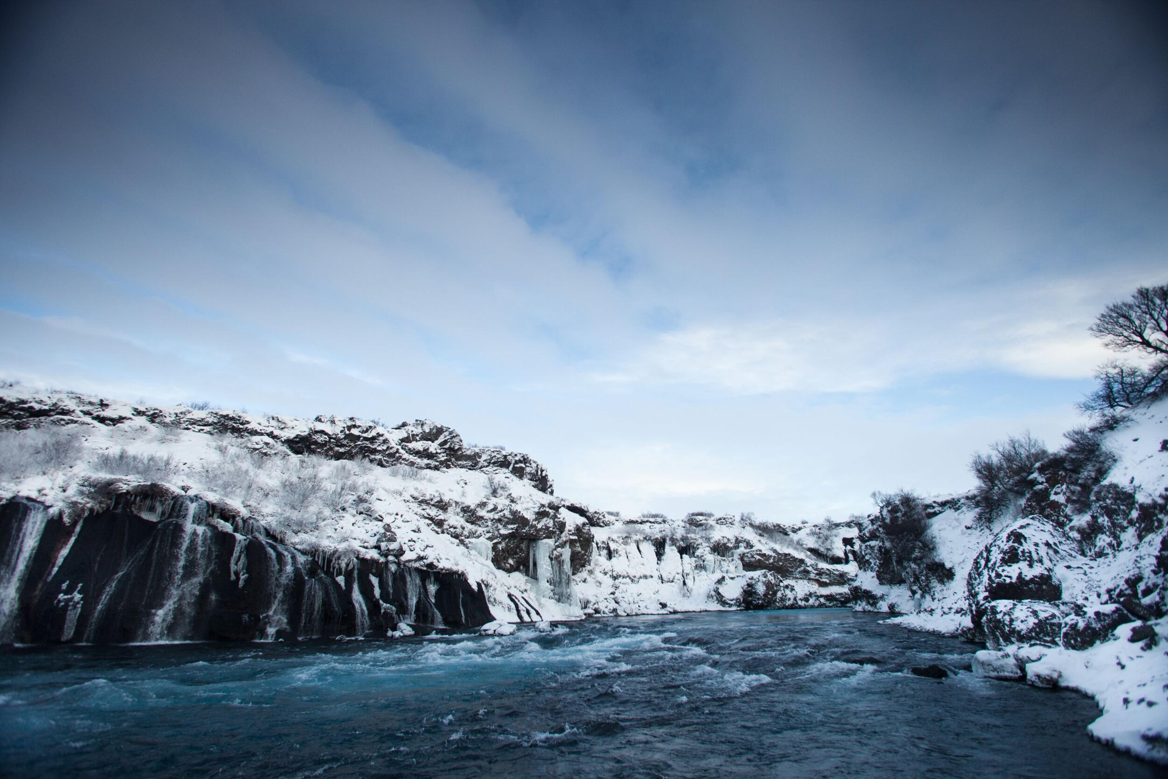 Hraunfossar waterfalls during winter, surrounded by snow-covered rocks and a vibrant blue river under a cloudy sky in Iceland.