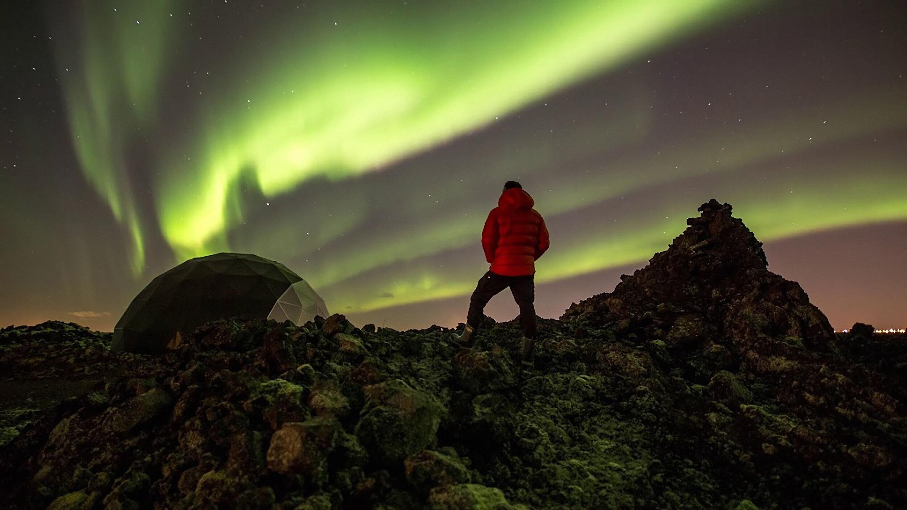 A visitor stands at Aurora Base Camp, Iceland, admire the northern lights as they illuminate the sky above a rugged, rocky landscape.