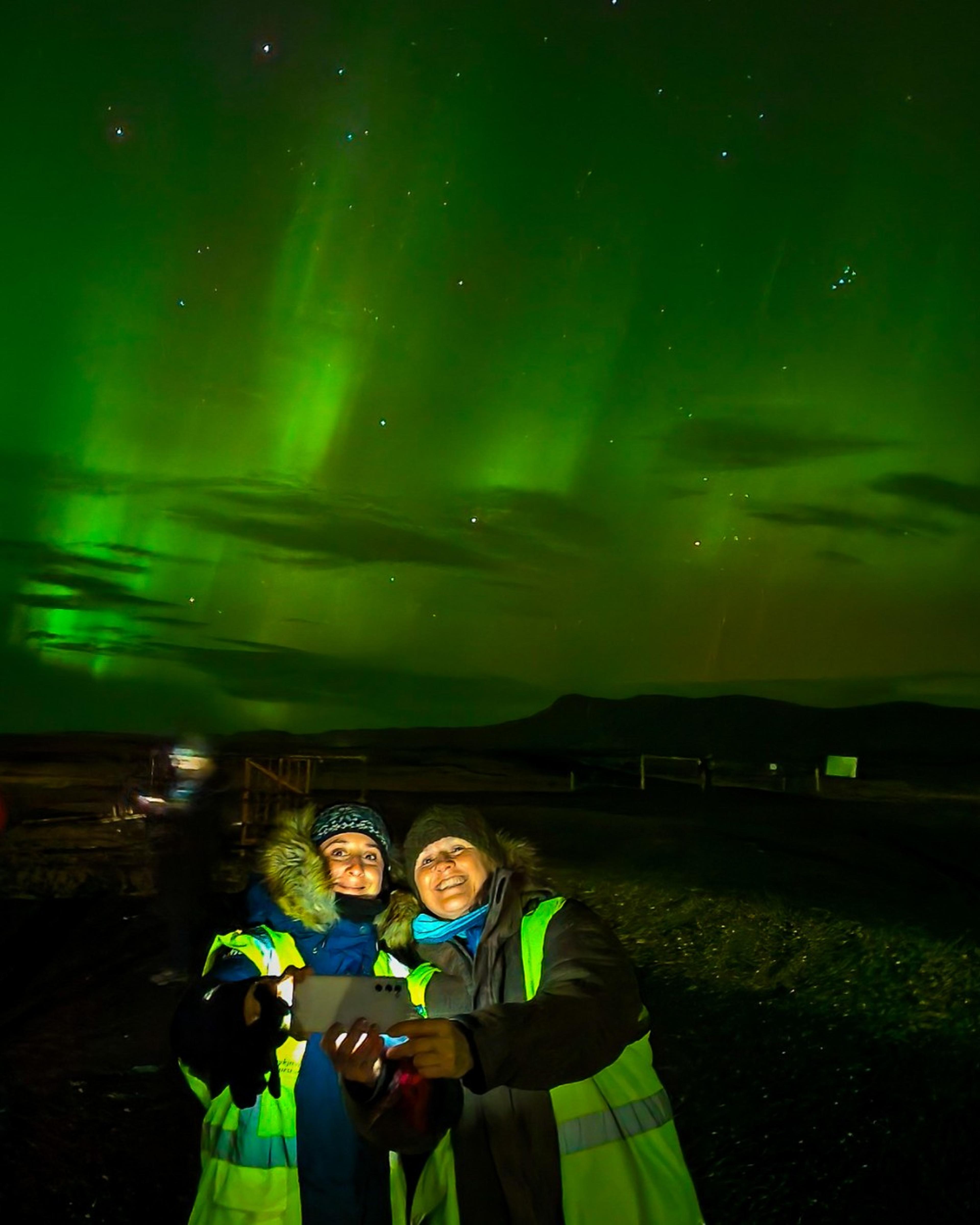 Two people wearing winter gear and reflective jackets take a selfie under a vibrant display of green Northern Lights in the night sky.