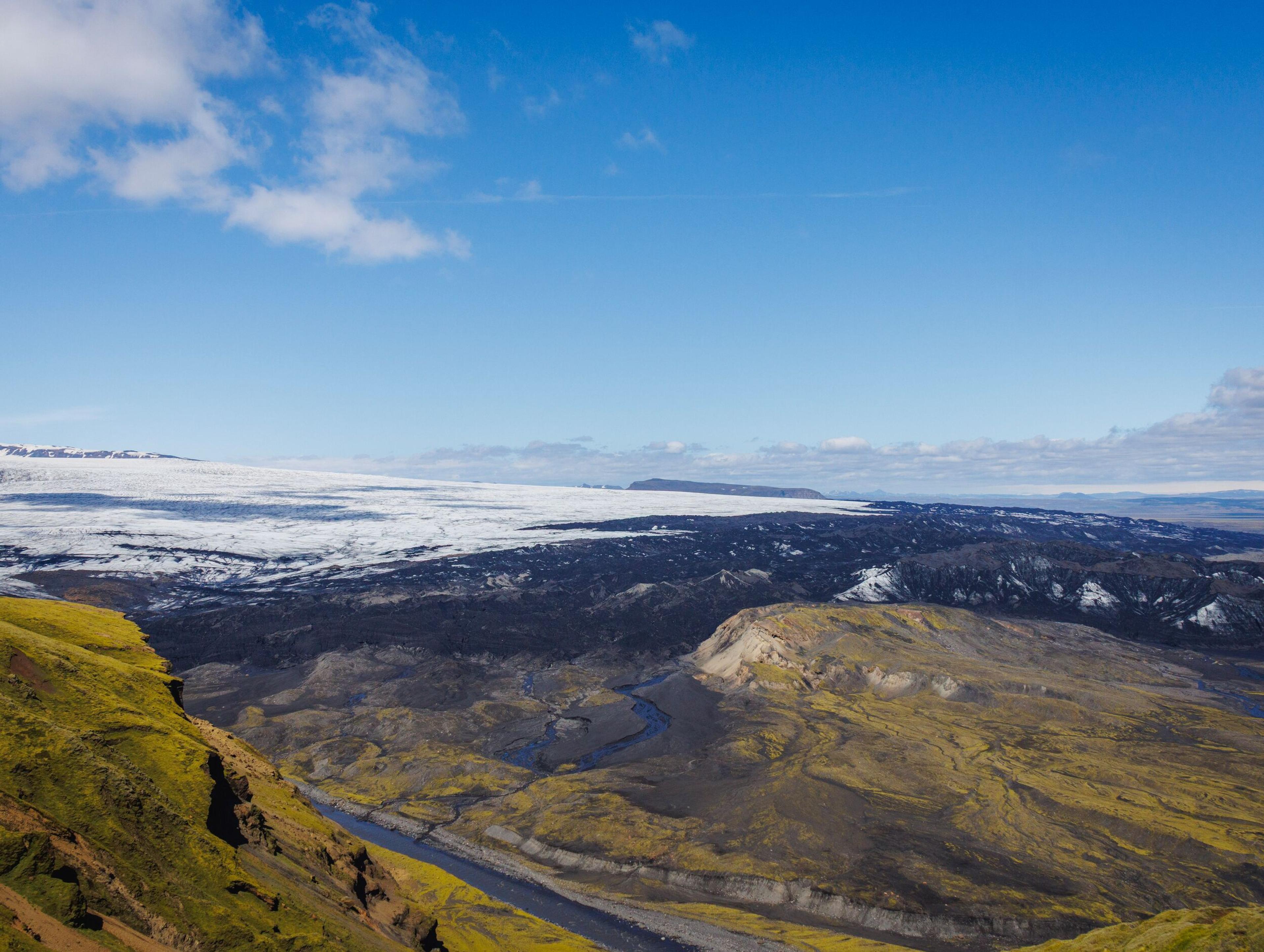 Panoramic view of a rugged landscape with a glacier, dark volcanic terrain, a winding river, and mossy hills under a blue sky.