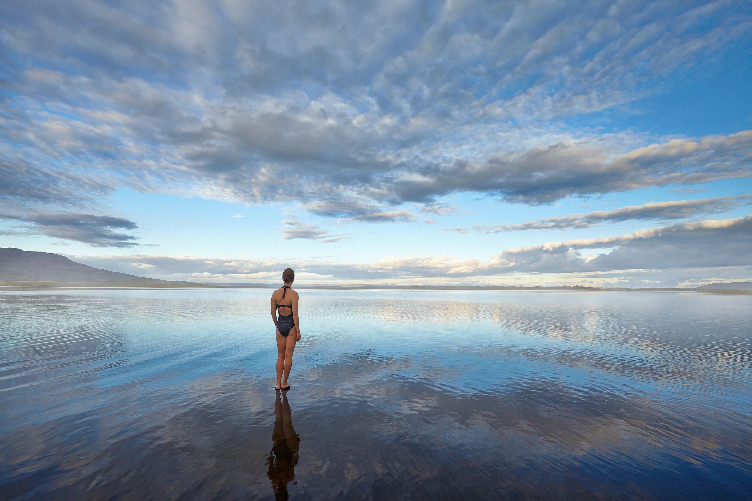 A woman stands in the shallow, reflective waters of Laugarvatn Fontana lake in Iceland, a serene geothermal bathing spot perfect for any Iceland itinerary.