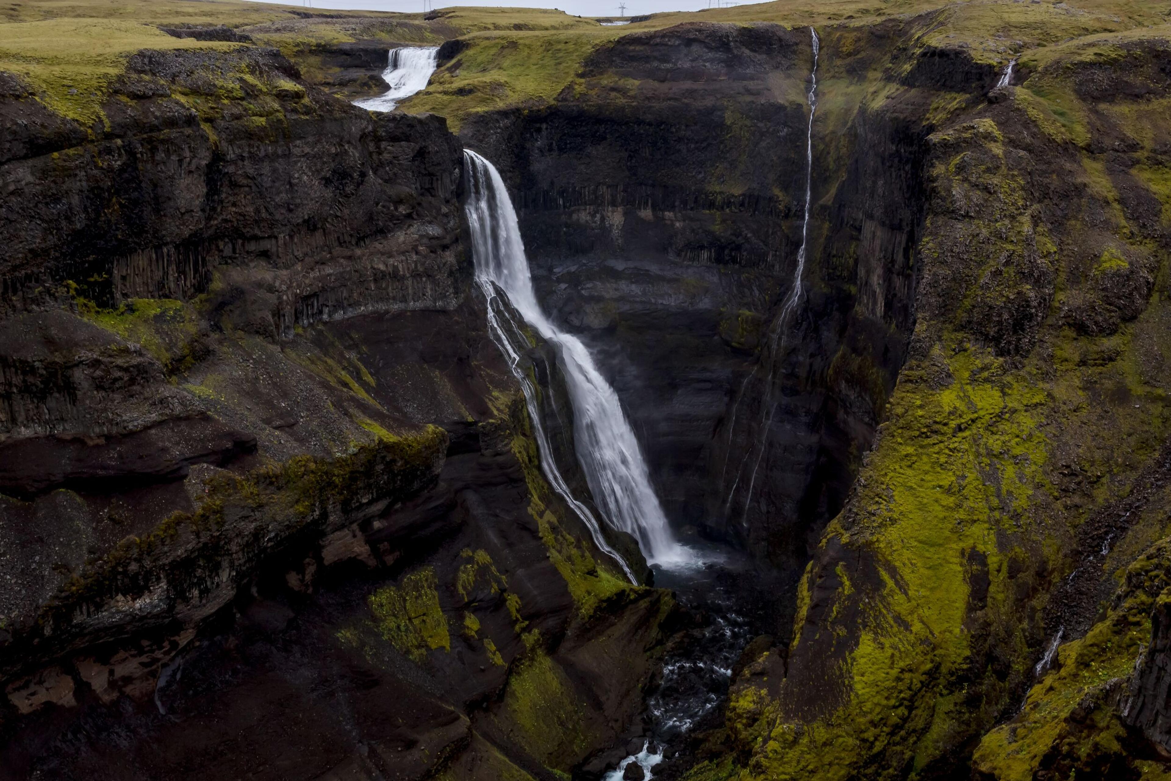 A deep canyon features a large waterfall, smaller streams, and vibrant green moss on its dark rock walls.