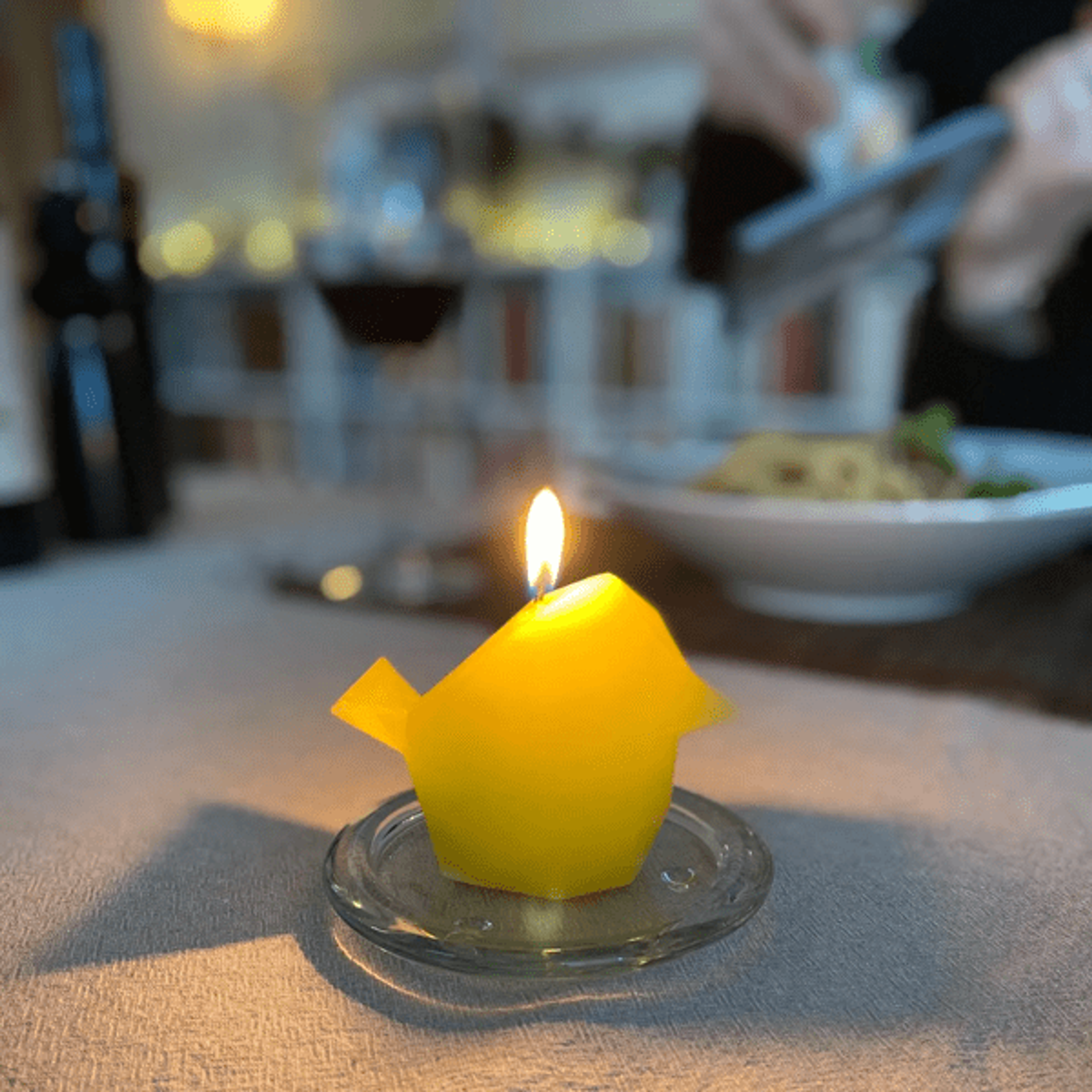 A lit yellow origami bird candle on a glass plate, with a blurred background of a dinner table scene.