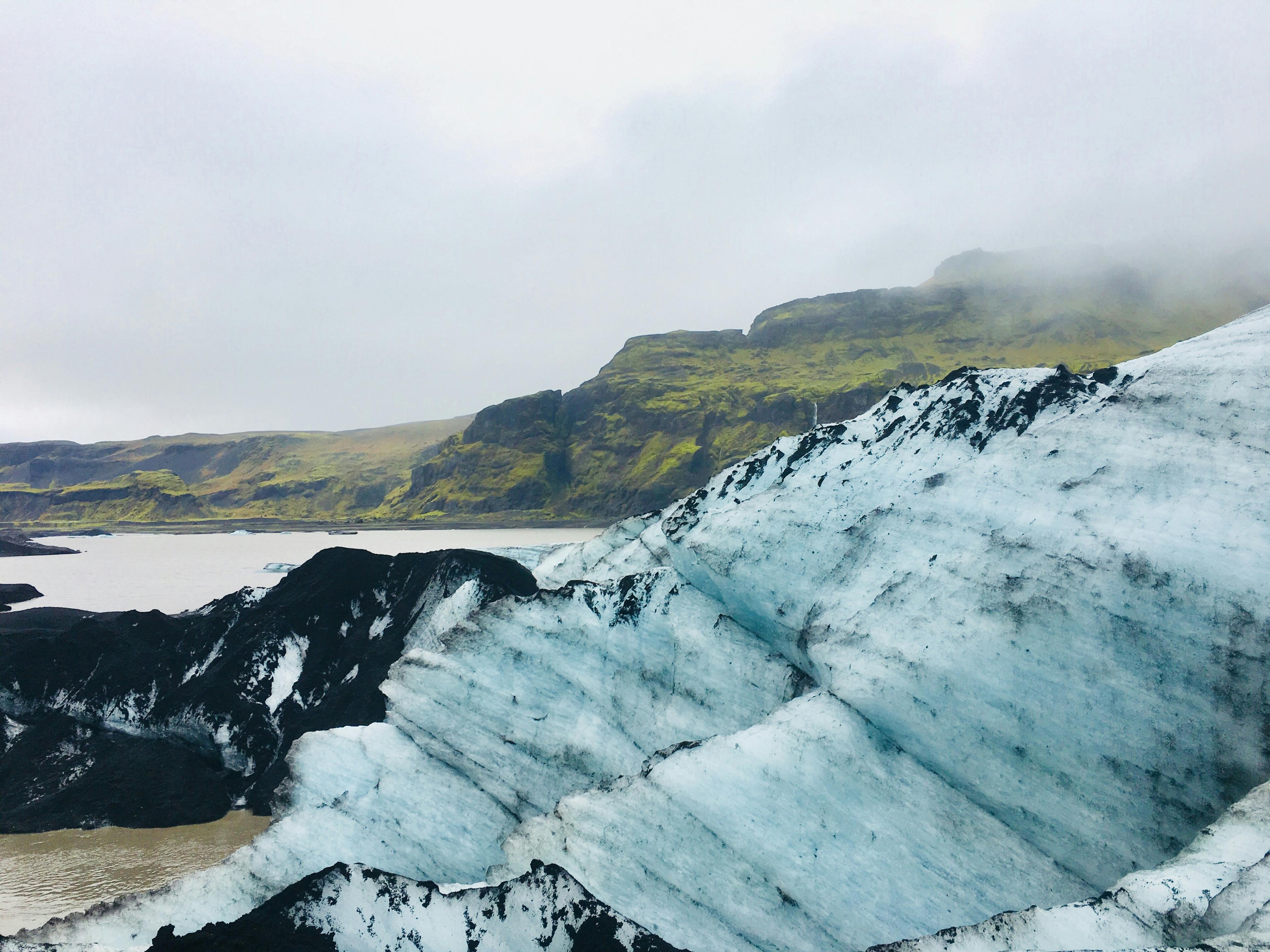 A striking contrast of blue ice and black volcanic ash on Sólheimajökull glacier, with moss-covered hills in the background. A stunning sight along the Sólheimajökull glacier hike in Iceland.