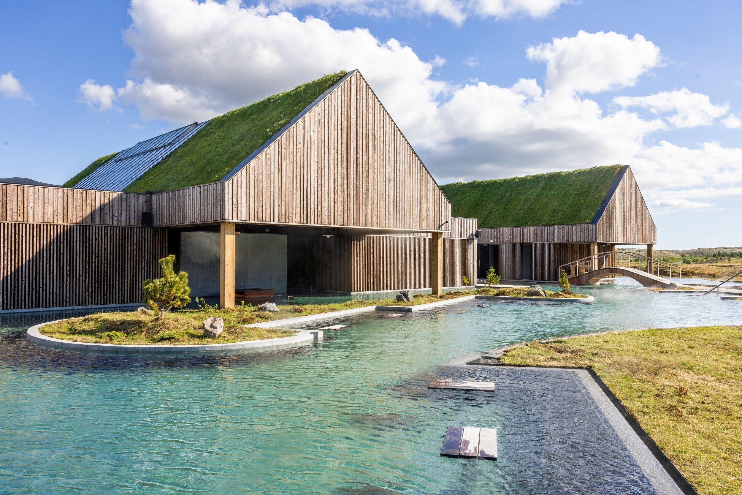 Modern spa buildings with green roofs and wooden siding next to light blue thermal pools.