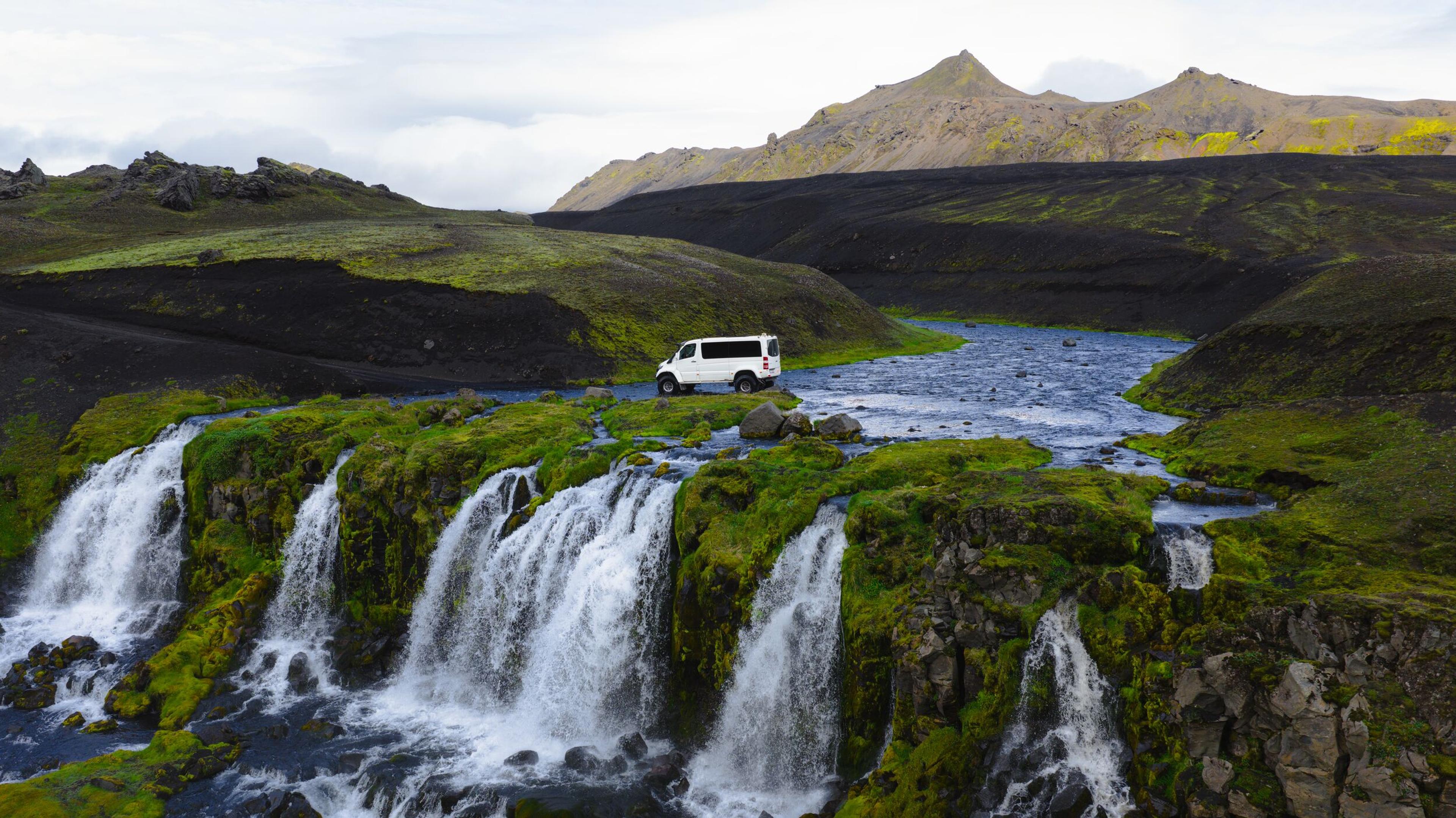 A white van parked by a river with multiple waterfalls, surrounded by green moss and black volcanic hills.