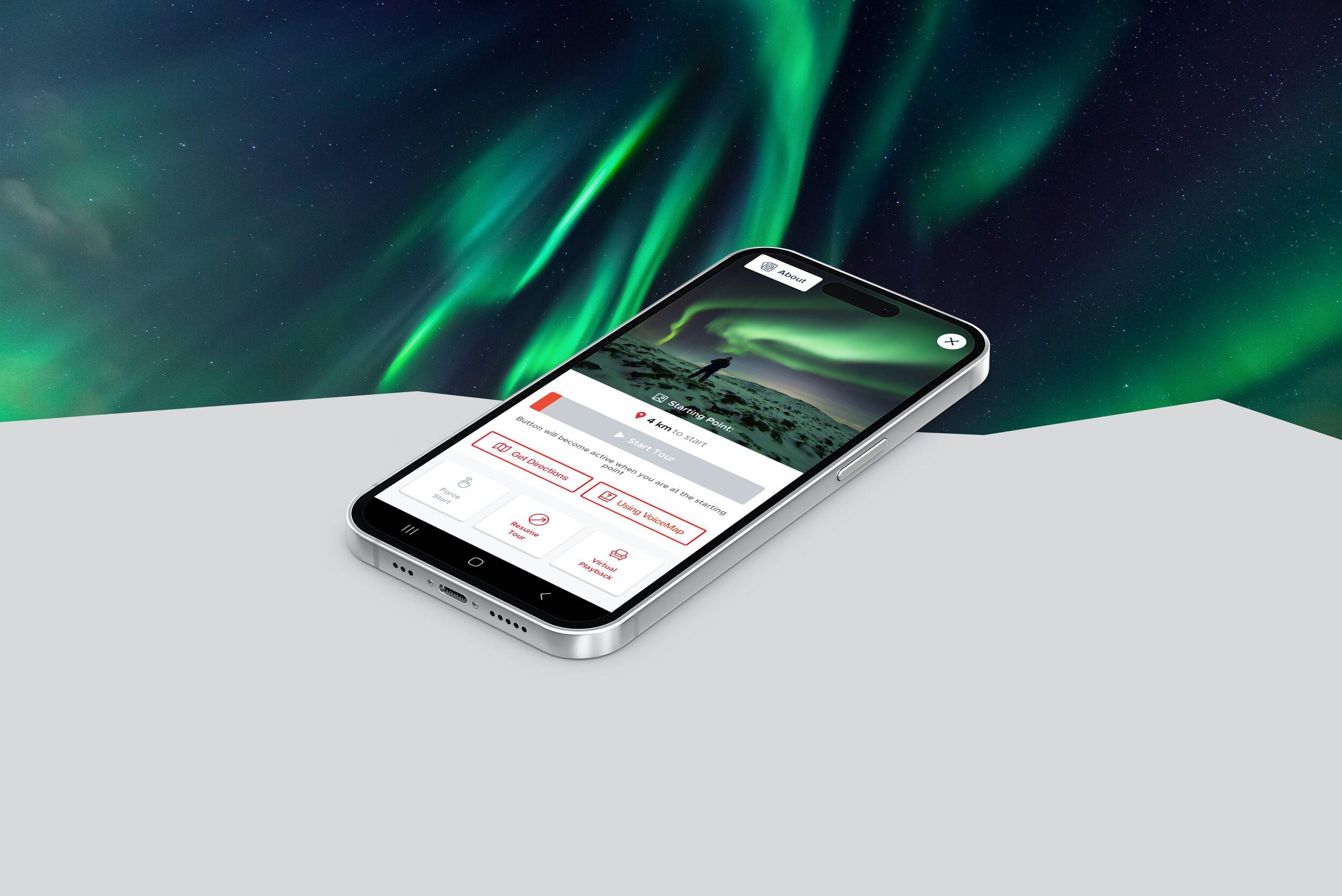 Black smartphone displaying VoipceMap audio guide app, set against a dramatic backdrop of the Northern Lights (Aurora Borealis) swirling in green hues above the horizon.
