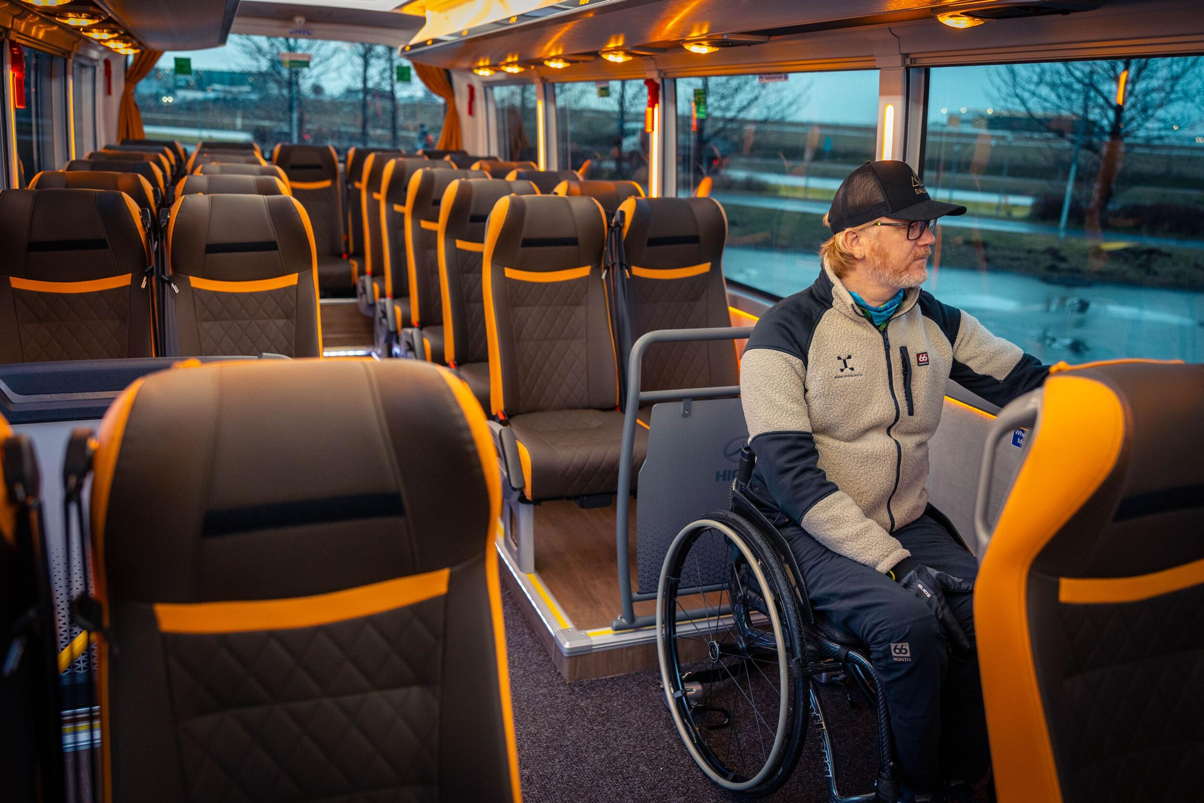 A man in a wheelchair sits inside a modern Flybus with black and orange seats, looking out the window.