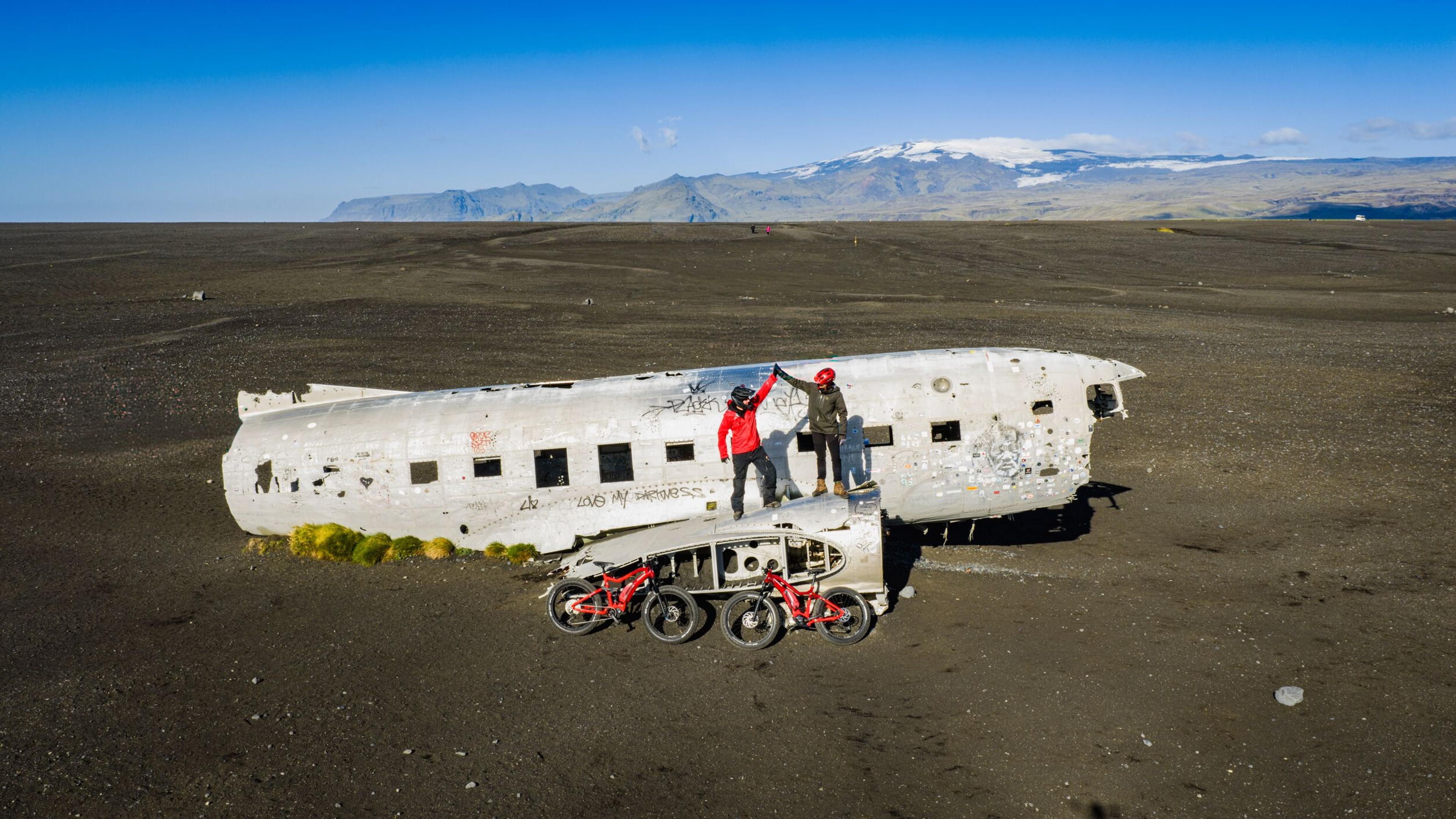 Two people high-five atop a multi-wheeled off-road vehicle next to a plane wreck fuselage on a dark plain, with snow-capped mountains in the background.