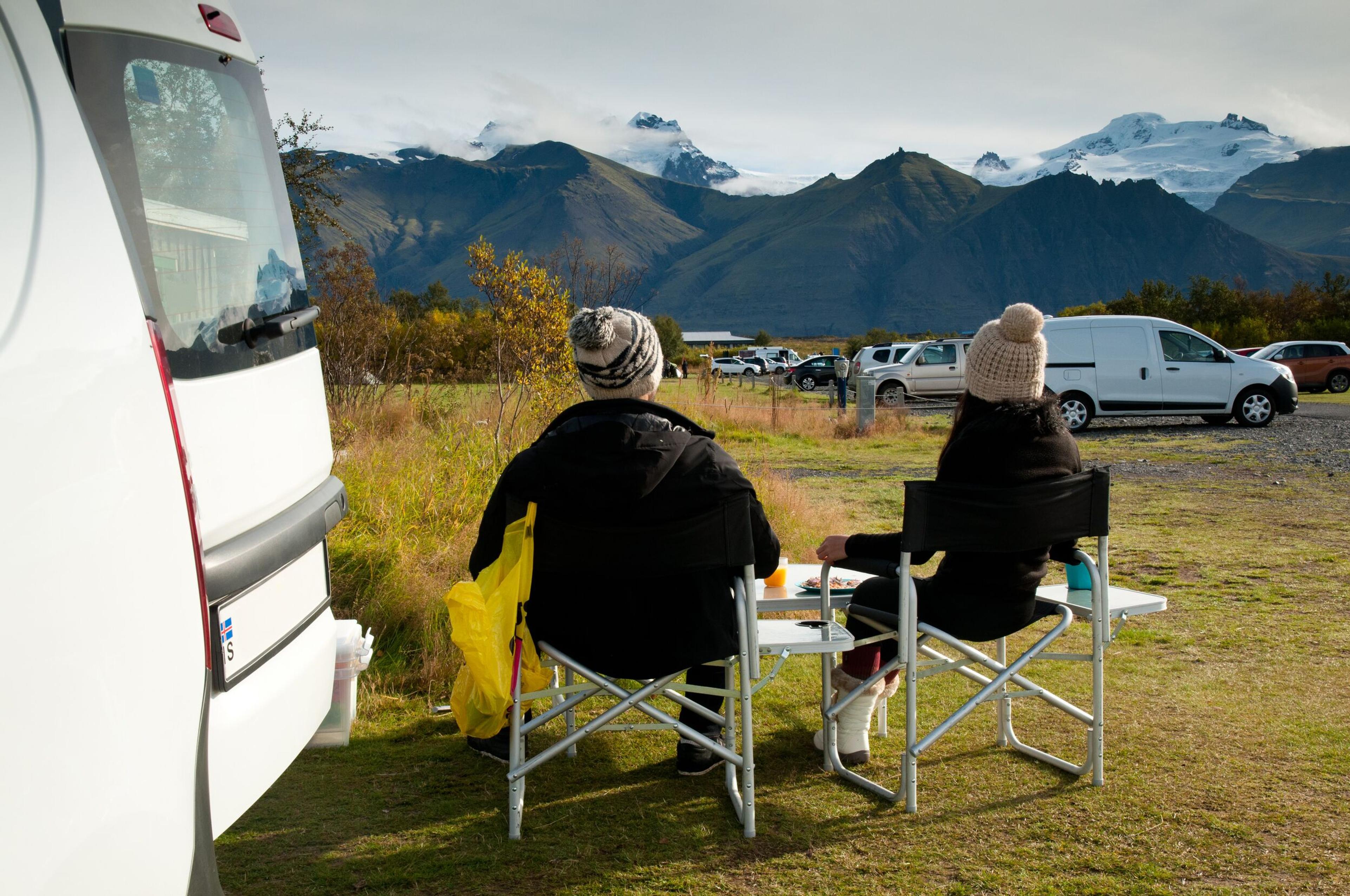 Two people seated in camping chairs beside a vehicle, enjoying a view of distant mountains with snow-capped peaks under a partly cloudy sky.