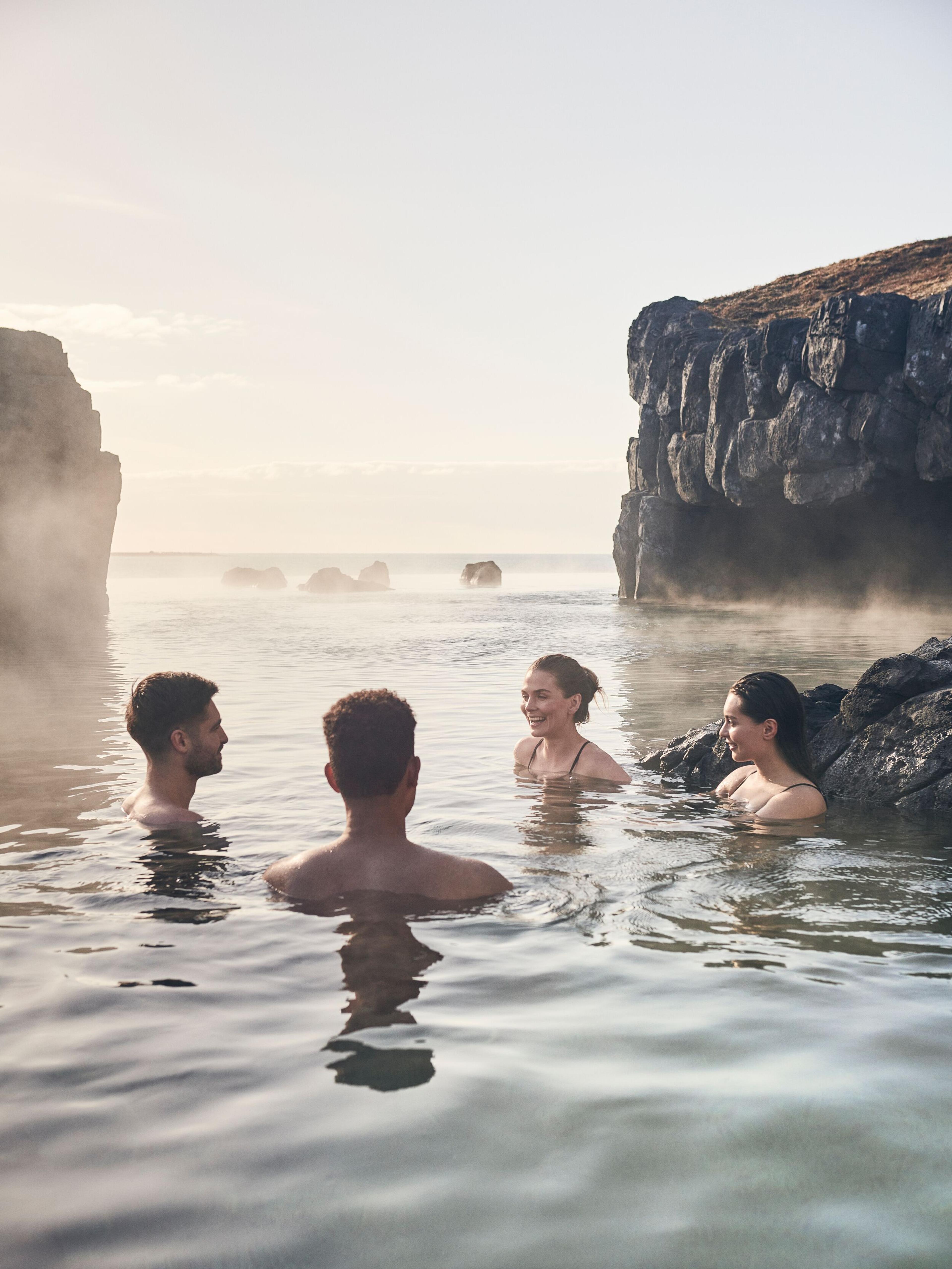 A group of friends enjoy a warm, convivial soak in the geothermal waters surrounded by the natural rocky landscape softened by the hazy mist of the hot spring.