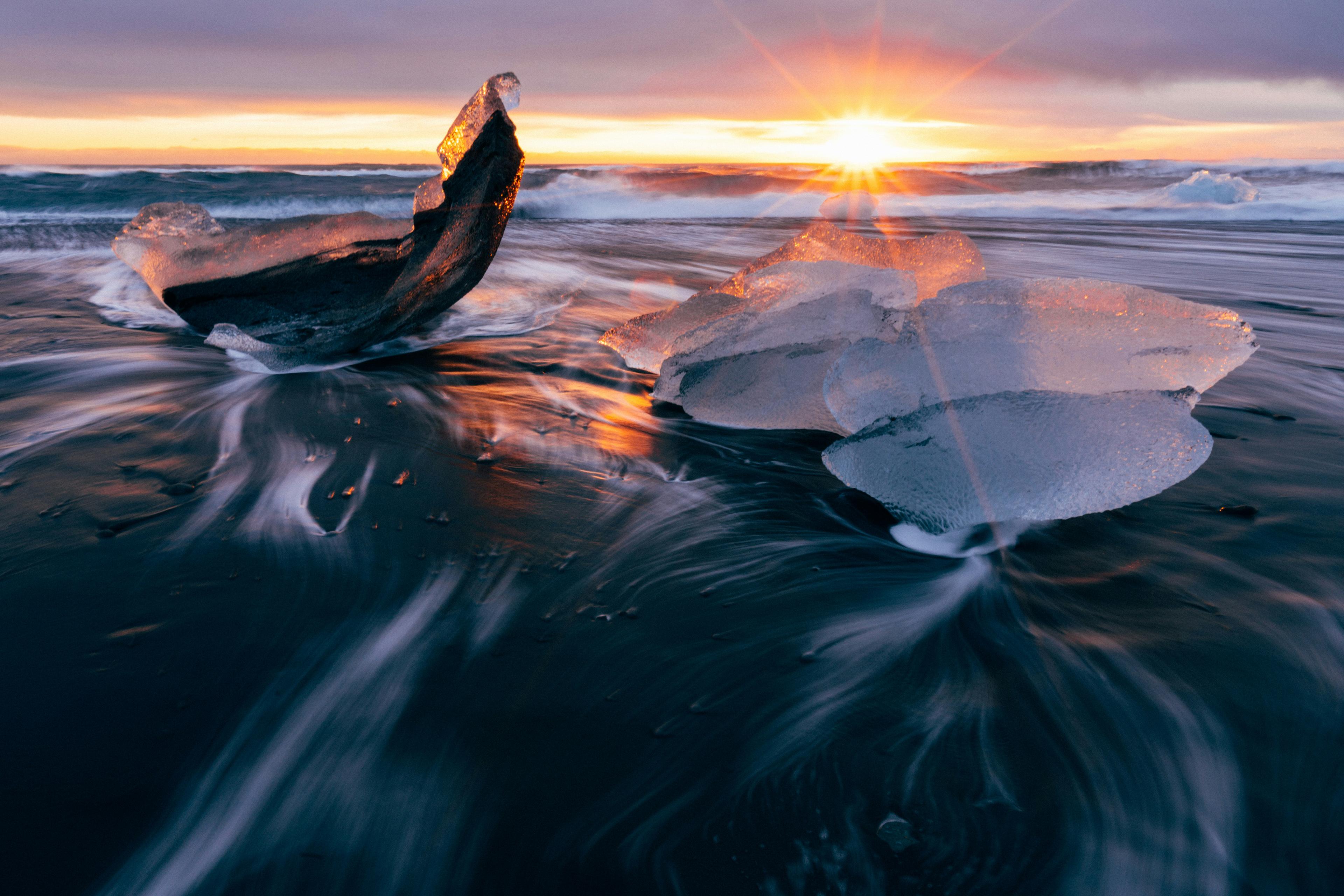 Sunrise over a black sand beach with translucent ice chunks scattered along the shore, waves lapping around them, creating patterns in the water.