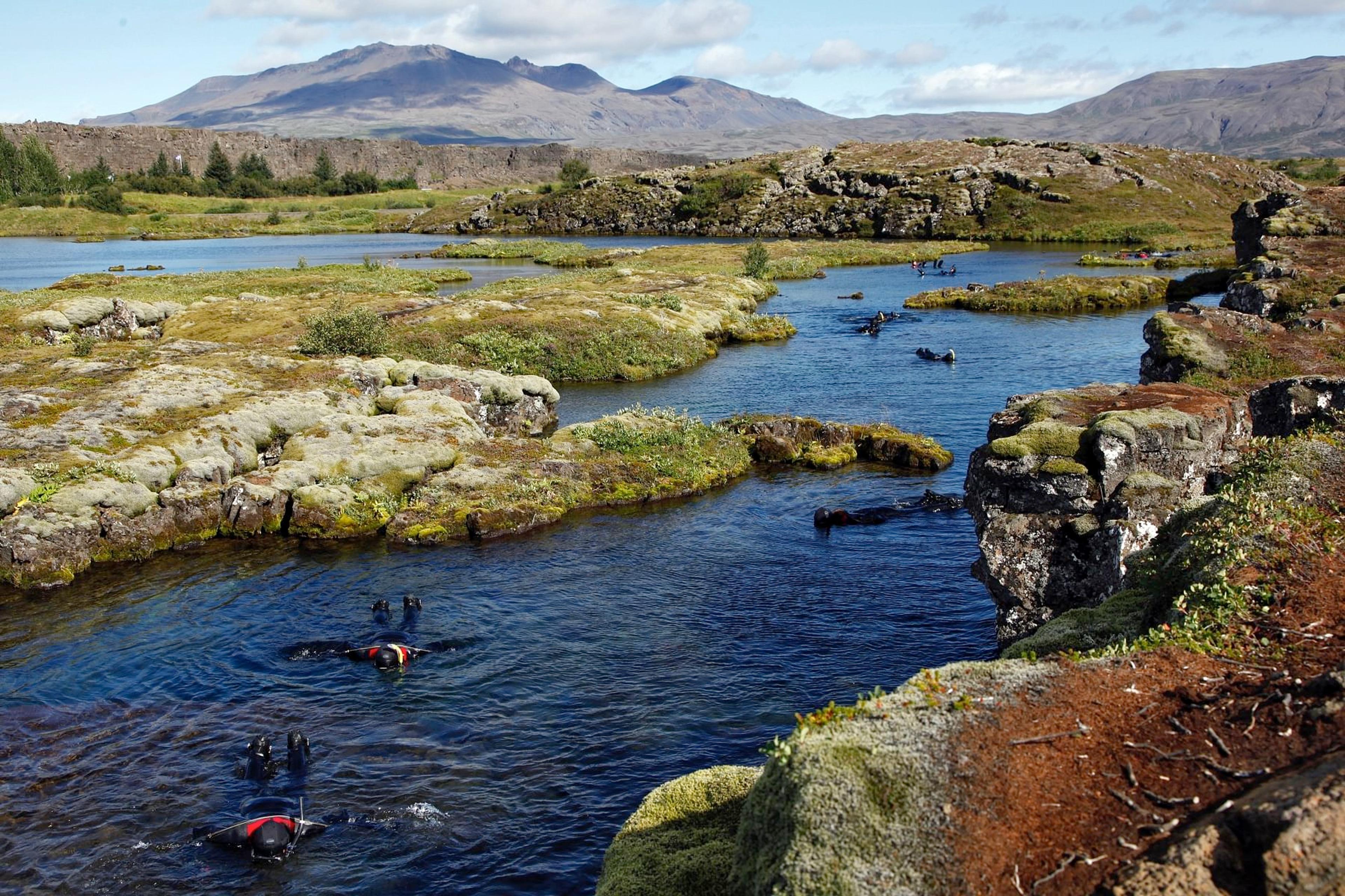 People in dry suits snorkel in a clear river flowing through a mossy, rocky landscape with distant mountains.