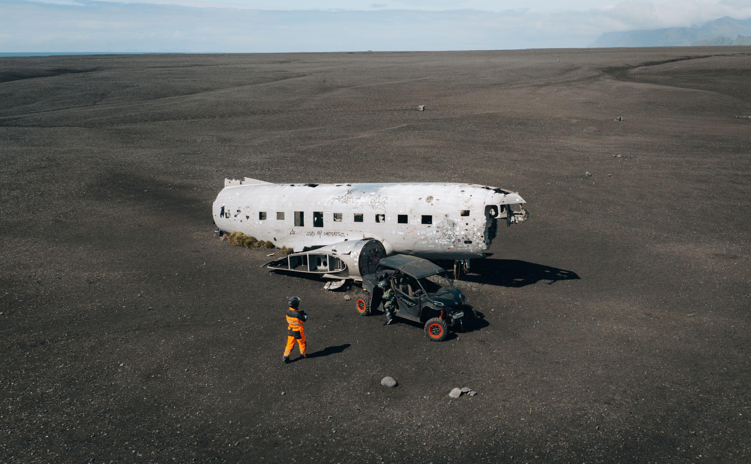 White airplane wreck on a black sand plain with a person in orange and an off-road vehicle.