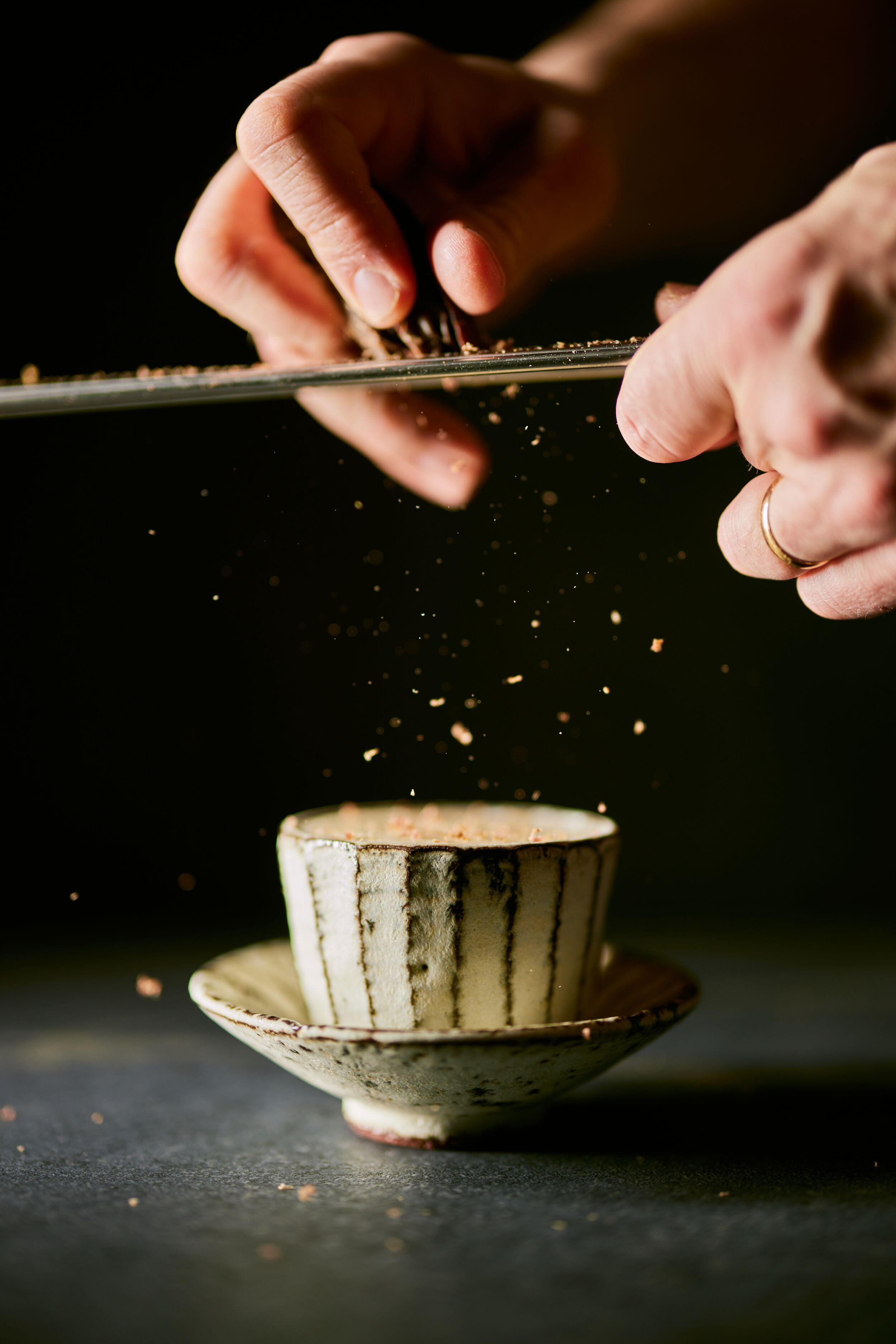 Hands grate a spice, creating falling particles, over a textured ceramic cup and saucer.