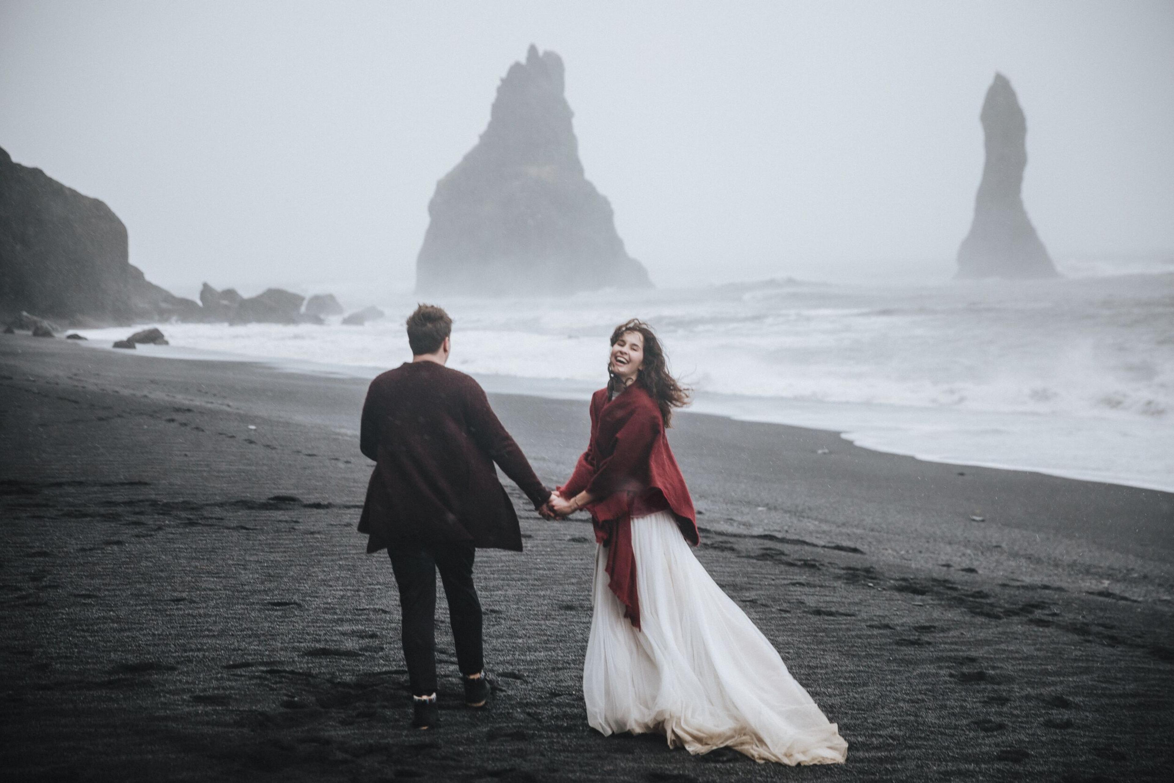 A couple holds hands on a black sand beach with sea stacks in the fog, the woman smiling back.