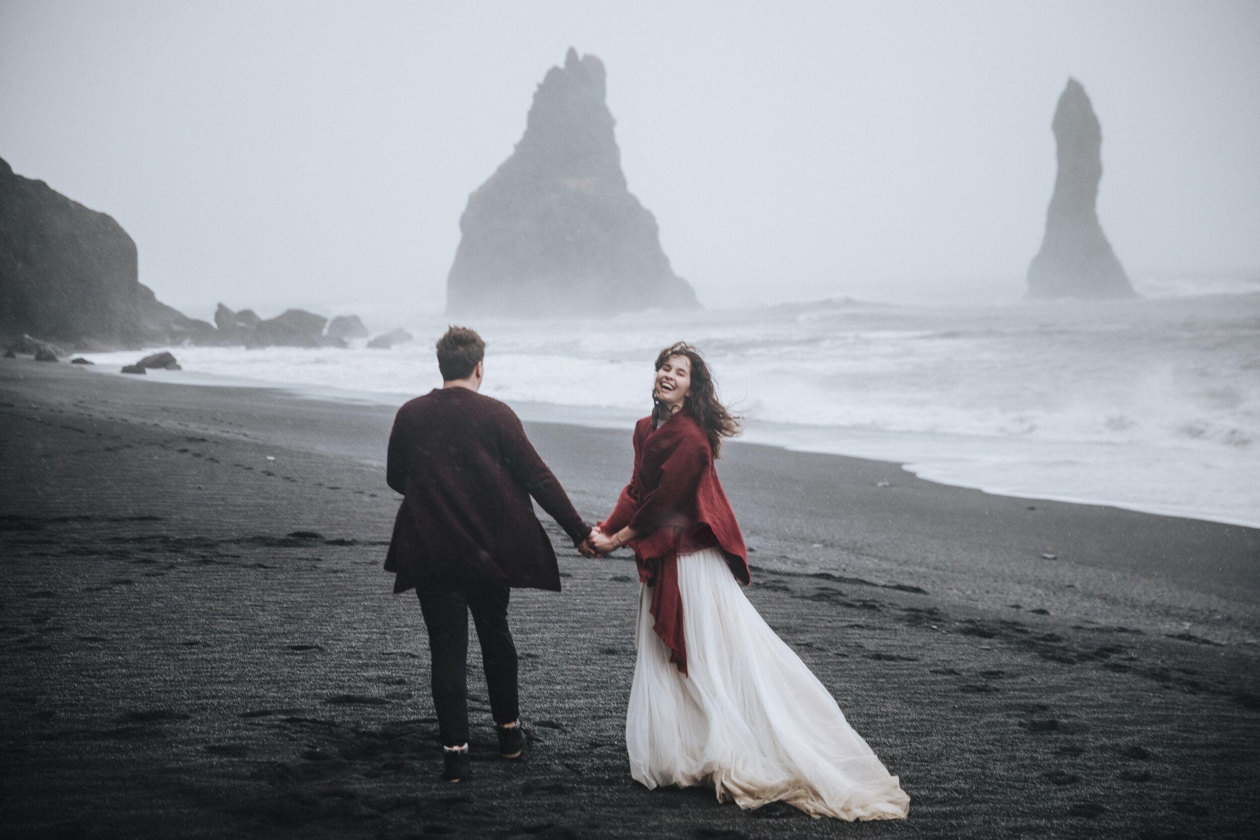 A couple holds hands on a black sand beach with sea stacks in the fog, the woman smiling back.