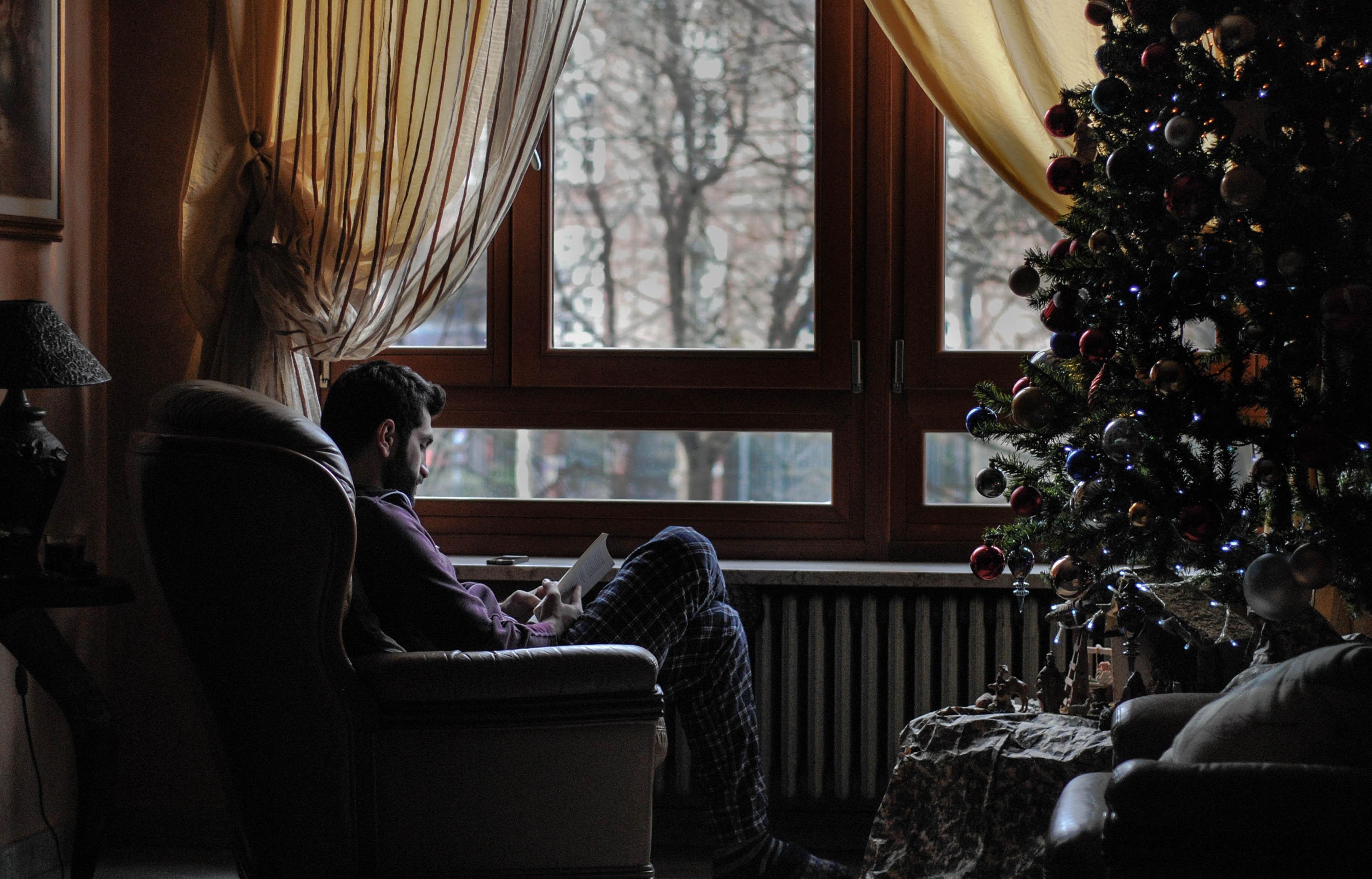 A man sitting in an armchair next to a Christmas tree and reading a book