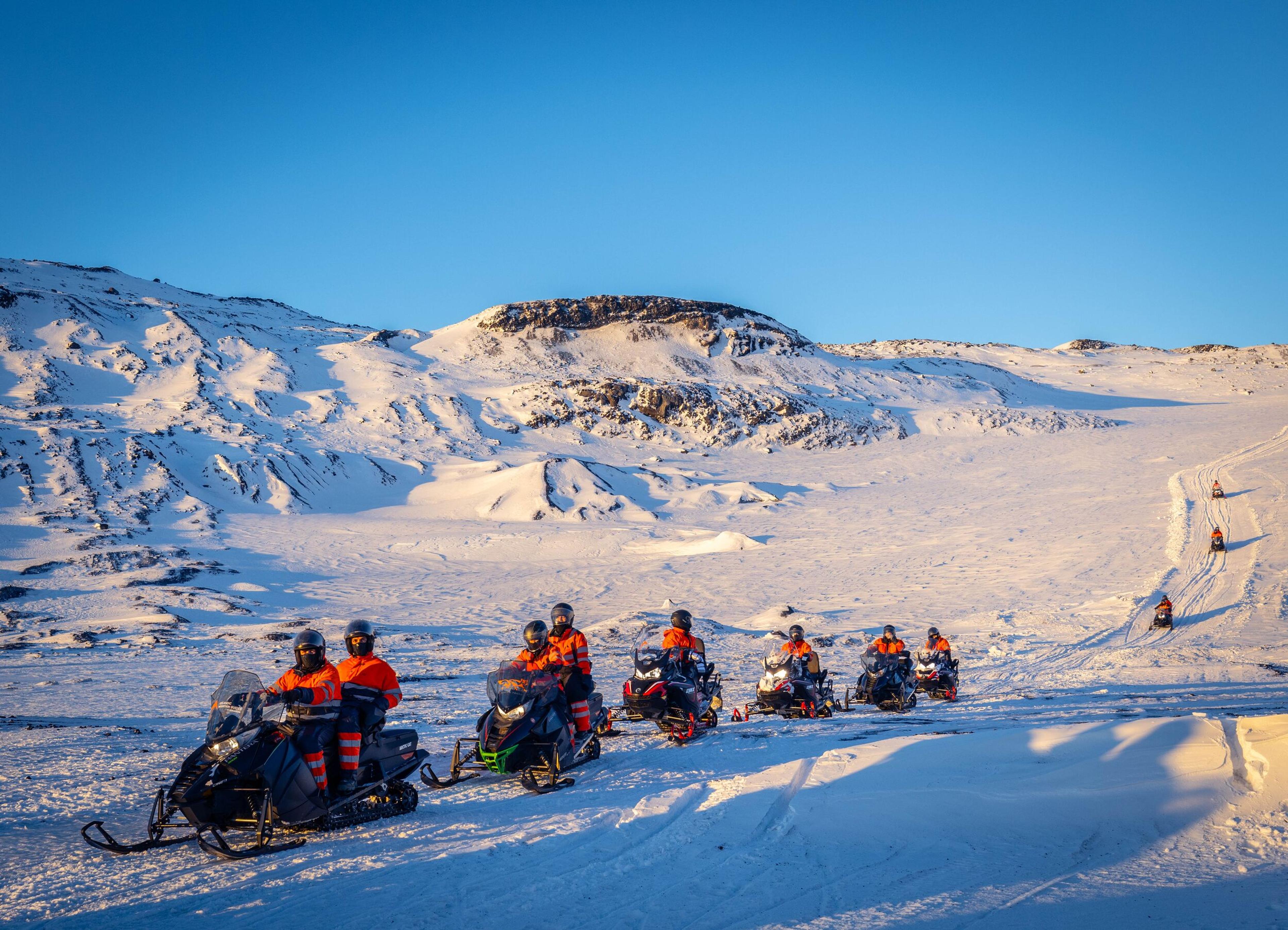 A group of adventurers on snowmobiles enjoy a bright day amidst a snowy landscape with rolling hills and a clear blue sky.