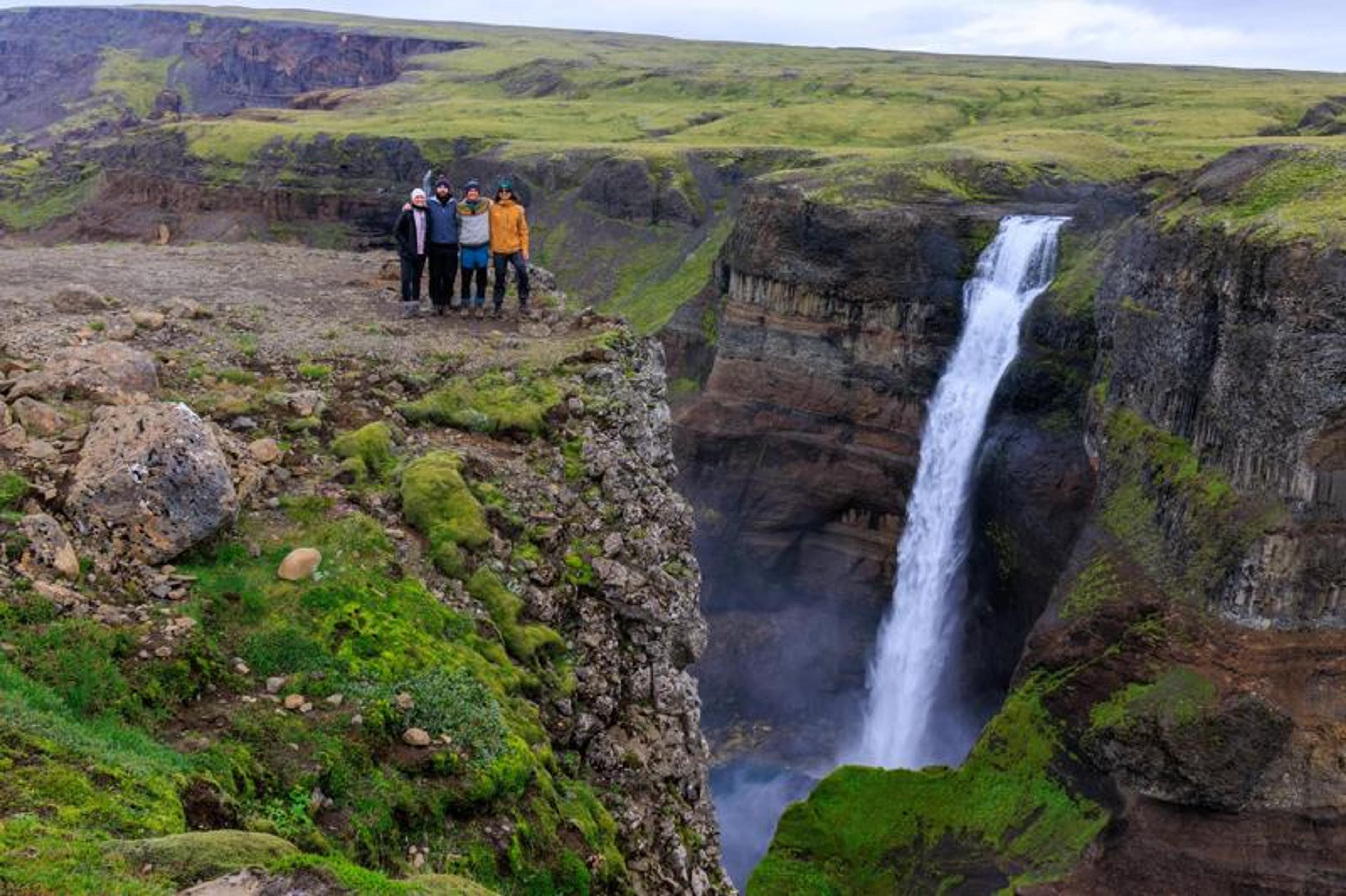 Five people stand on a rocky cliff beside a large waterfall cascading into a green canyon.