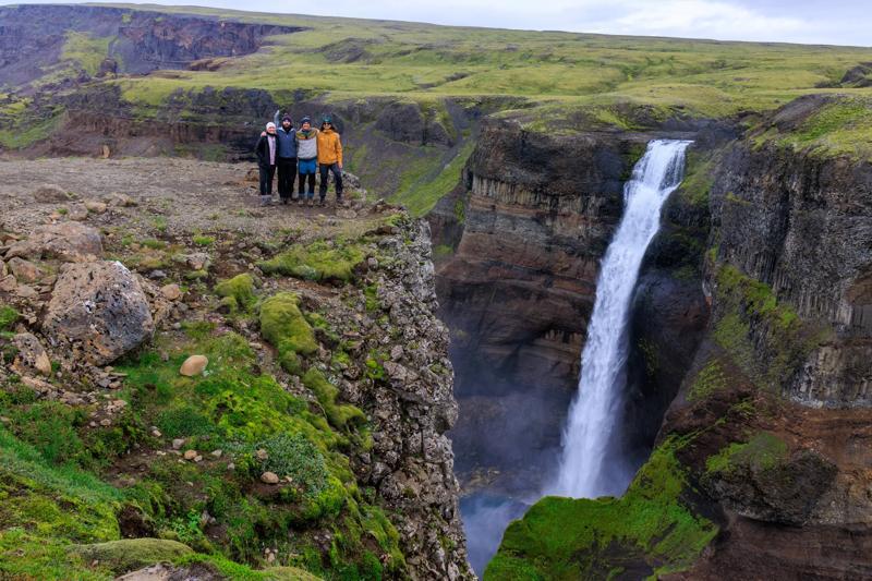 Five people stand on a rocky cliff beside a large waterfall cascading into a green canyon.