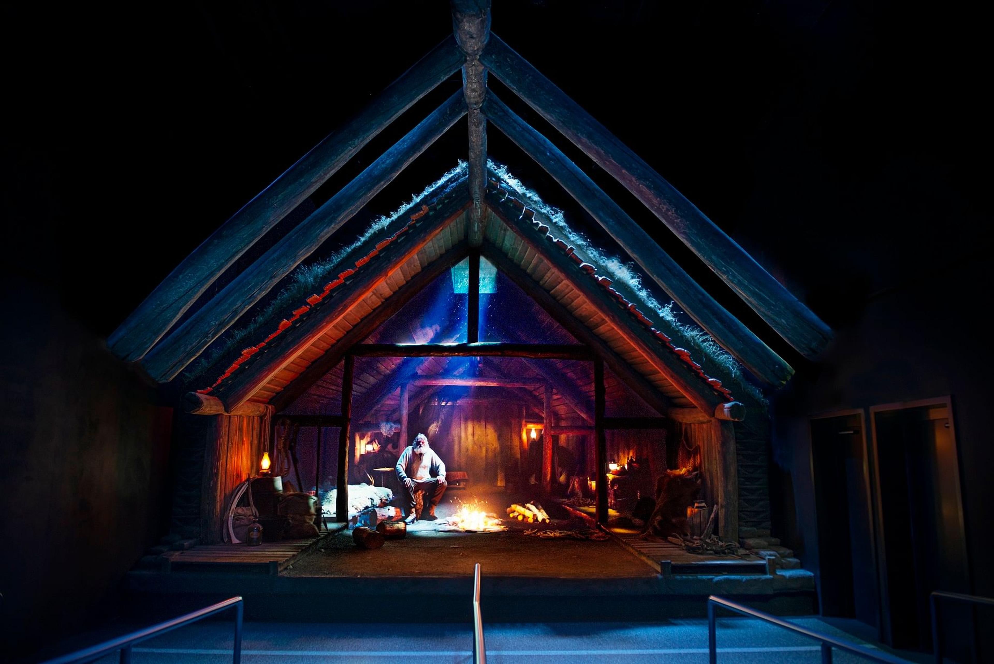 Museum exhibit of a rustic cabin interior with exposed beams, a figure by a fire, under orange and blue light.