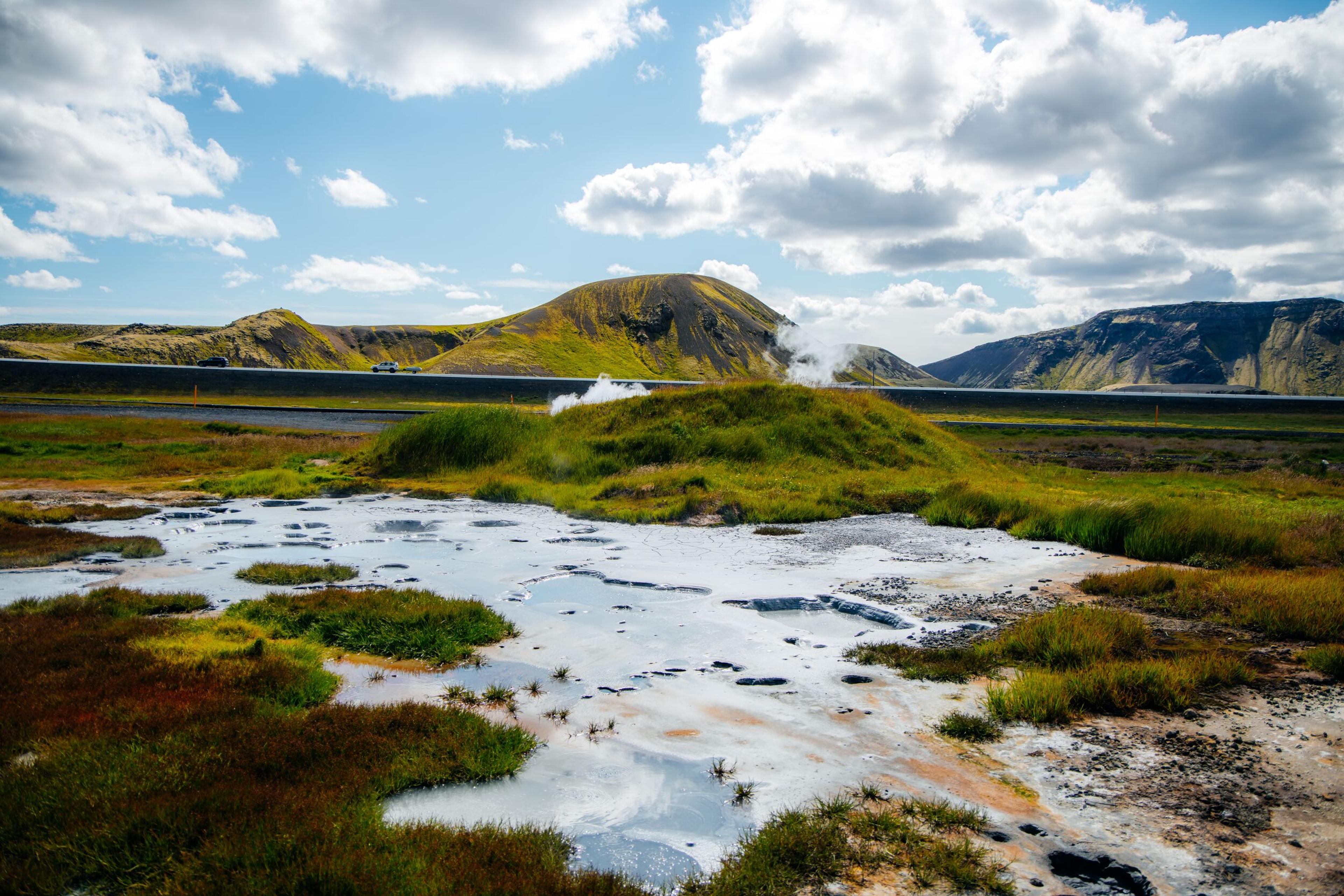 Geothermal landscape with steaming ground, colorful mineral pools, a road, and hills under a cloudy sky.