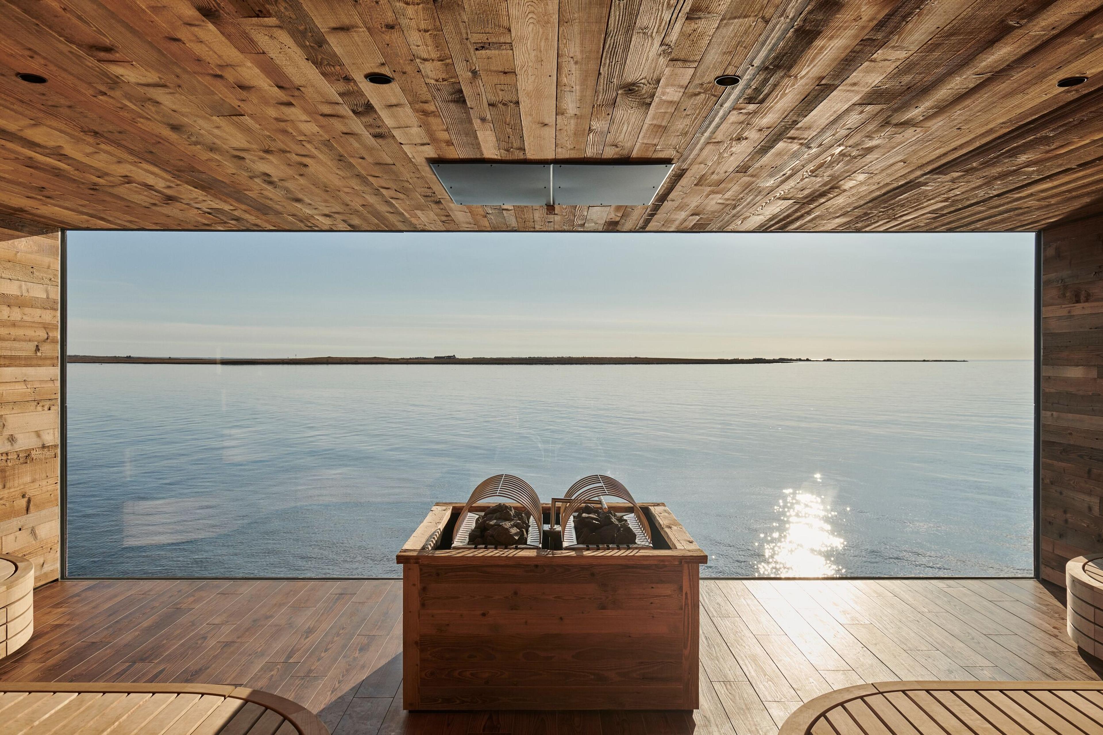 The interior of a sauna at Sky Lagoon with rustic wooden walls and ceiling, featuring a large window that offers a panoramic view of the calm sea and horizon, illuminated by the sun’s reflection on the water.