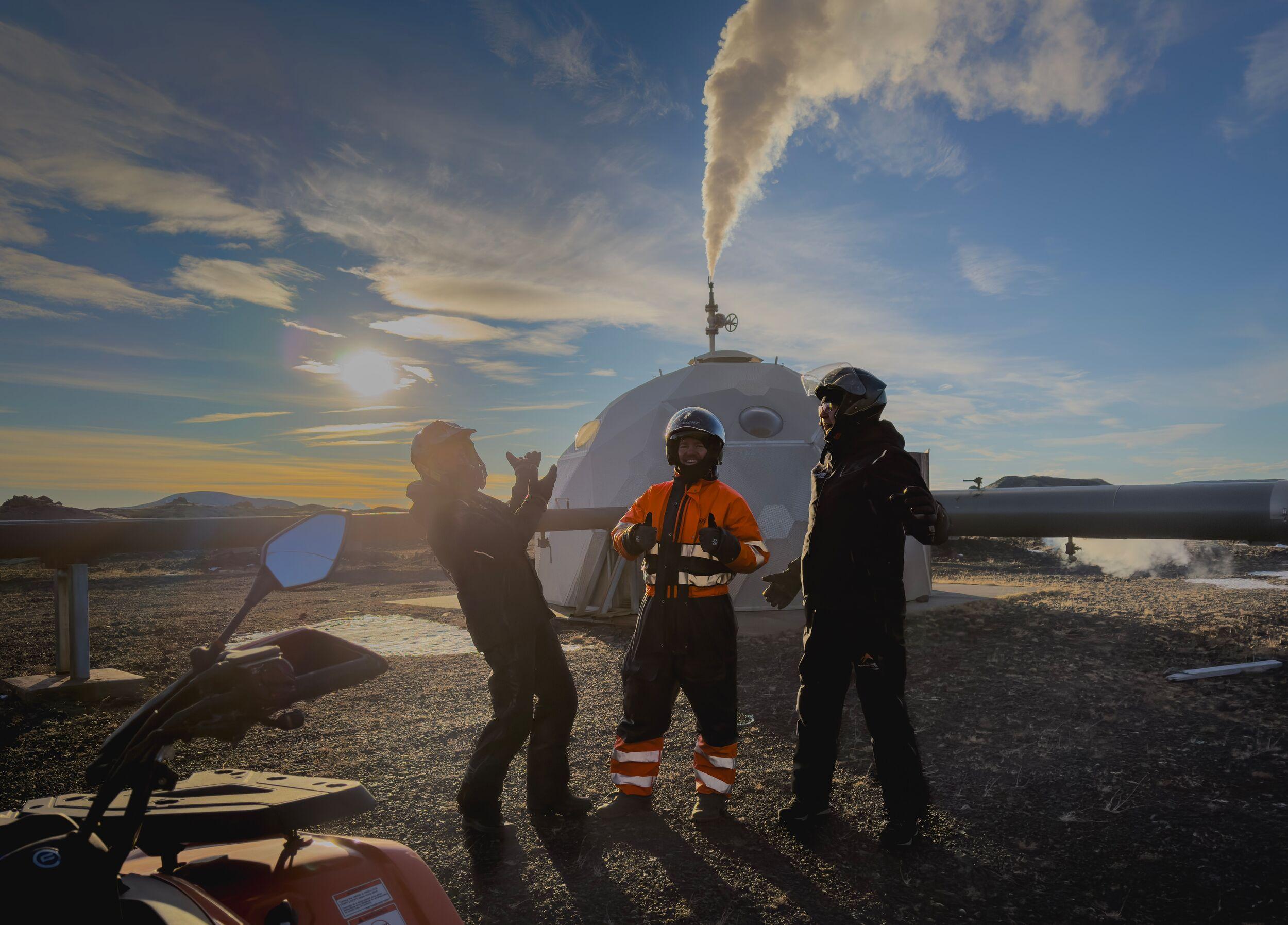 Three people in helmets and outdoor gear pose with an ATV at a geothermal site featuring a dome structure emitting steam, under a golden sunset.