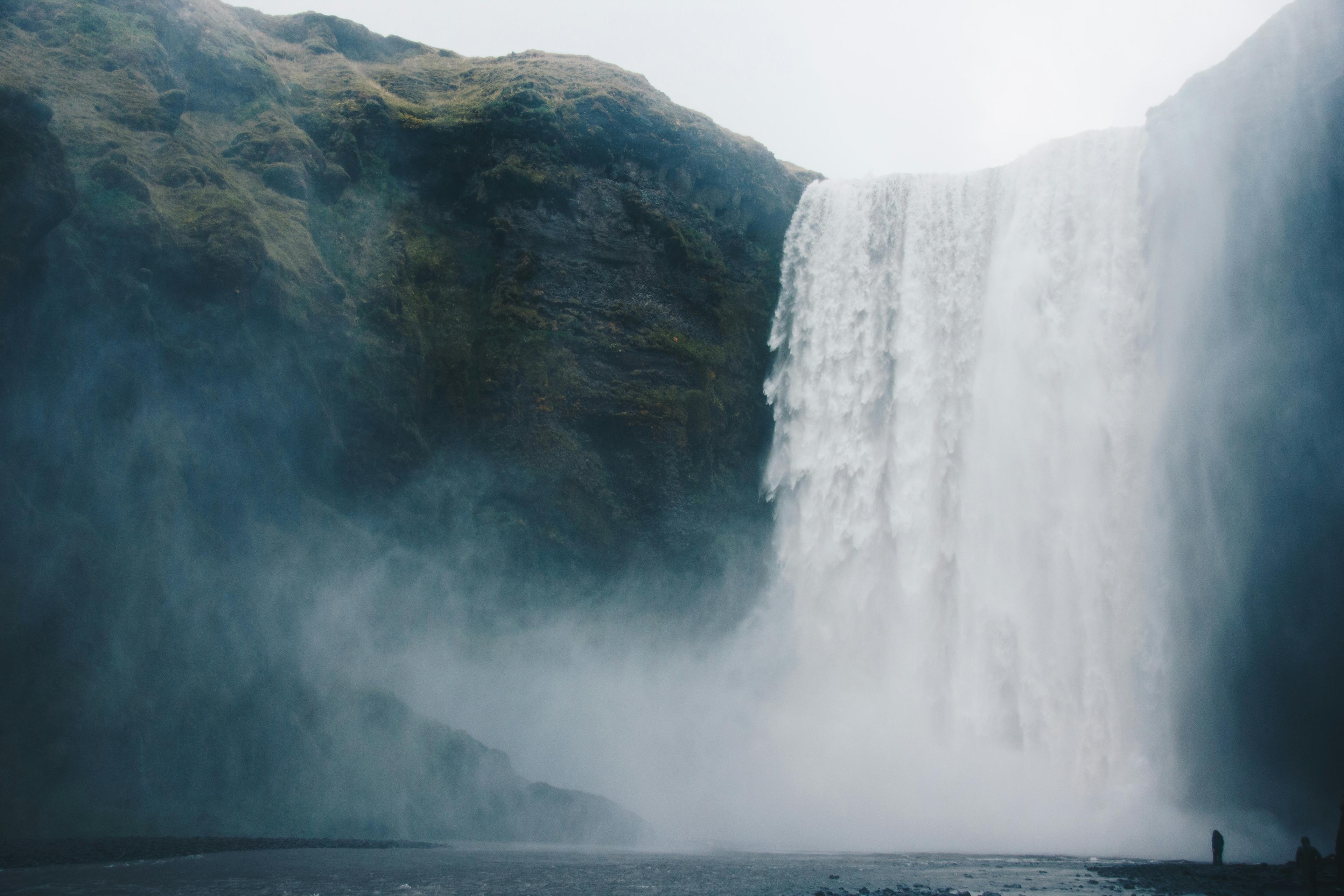 Skógafoss Waterfall in Iceland flowing down a moss-covered cliff, creating mist and with a person standing at the base for perspective.