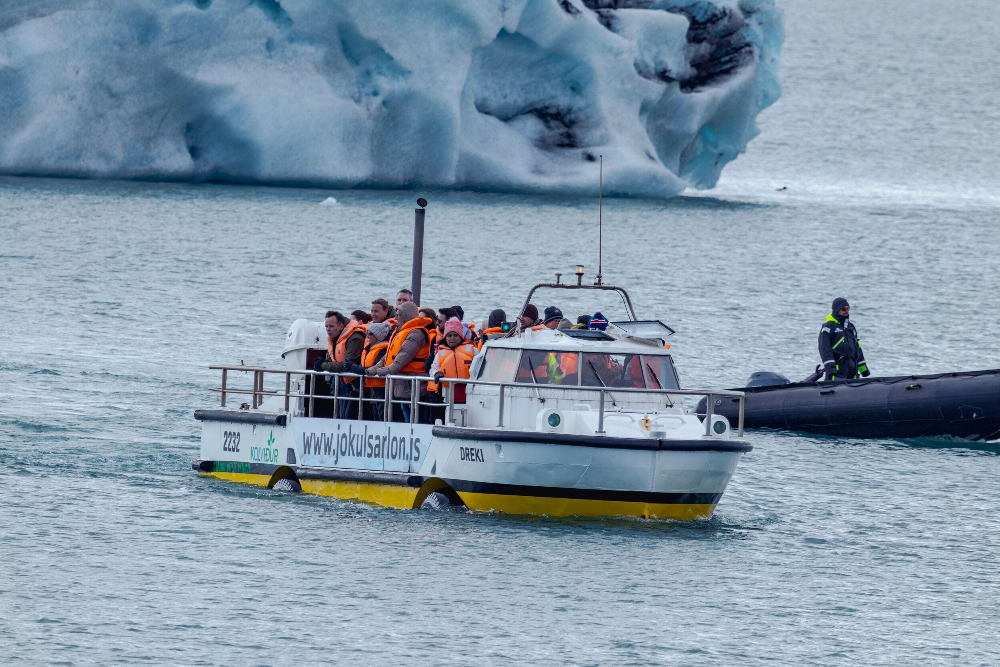 A tour boat filled with passengers in orange life vests cruises on a lake near a large iceberg, with a smaller inflatable boat nearby.