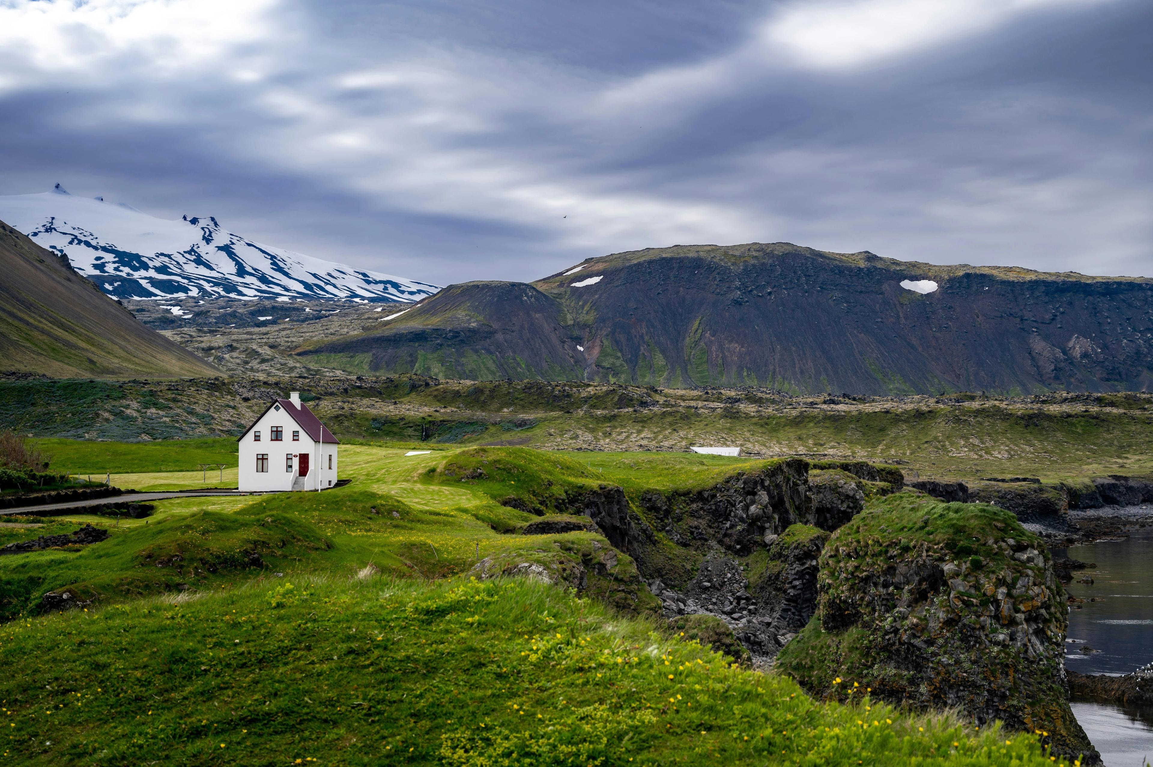 A white house with a red door stands in a lush green landscape with rocky cliffs and snow-capped mountains under an overcast sky.