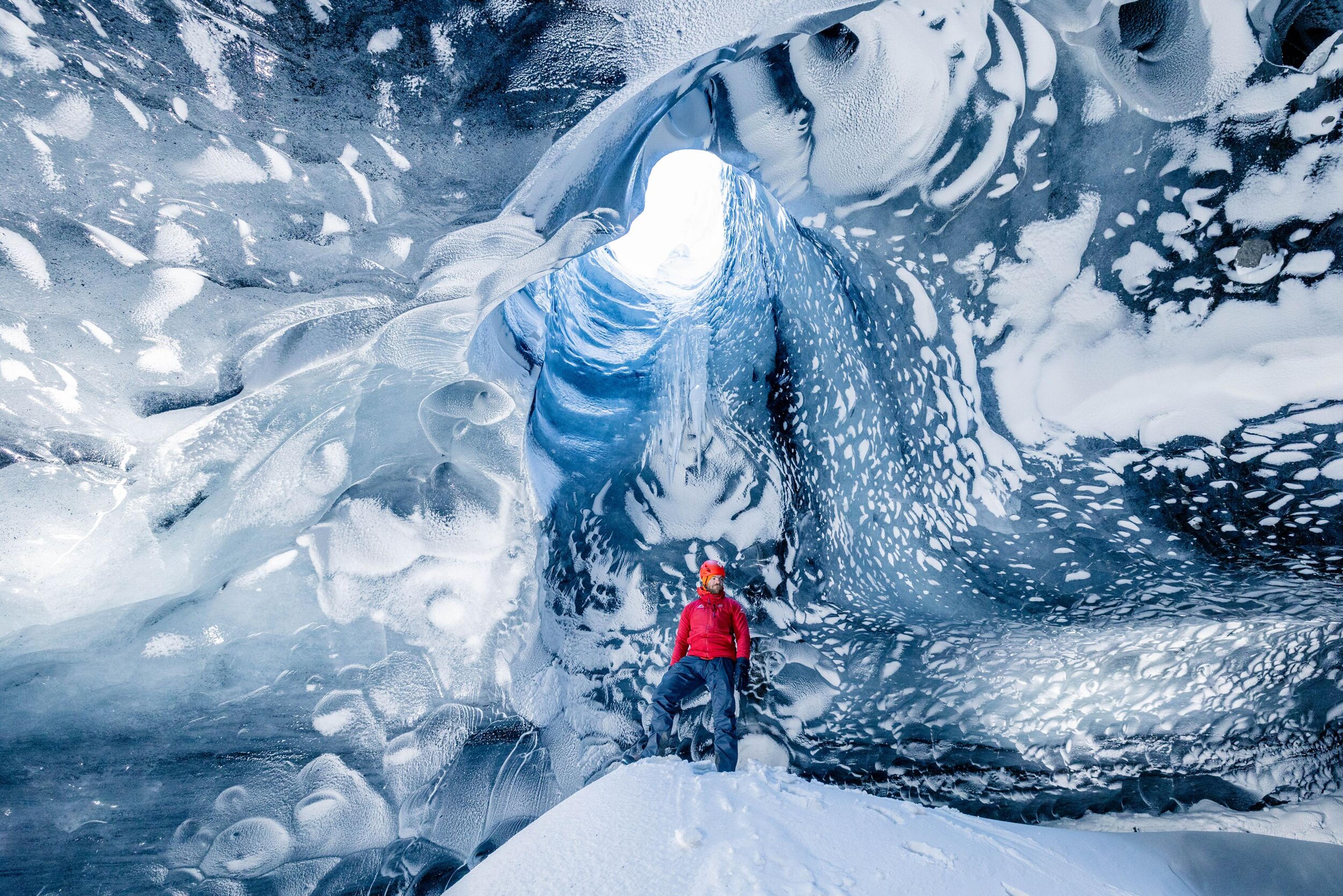 The view from inside a spacious ice cave looking outwards, with a person in a red jacket visible in the distance. The cave is characterized by its remarkable icy structures that hang from the ceiling and rise from the ground.