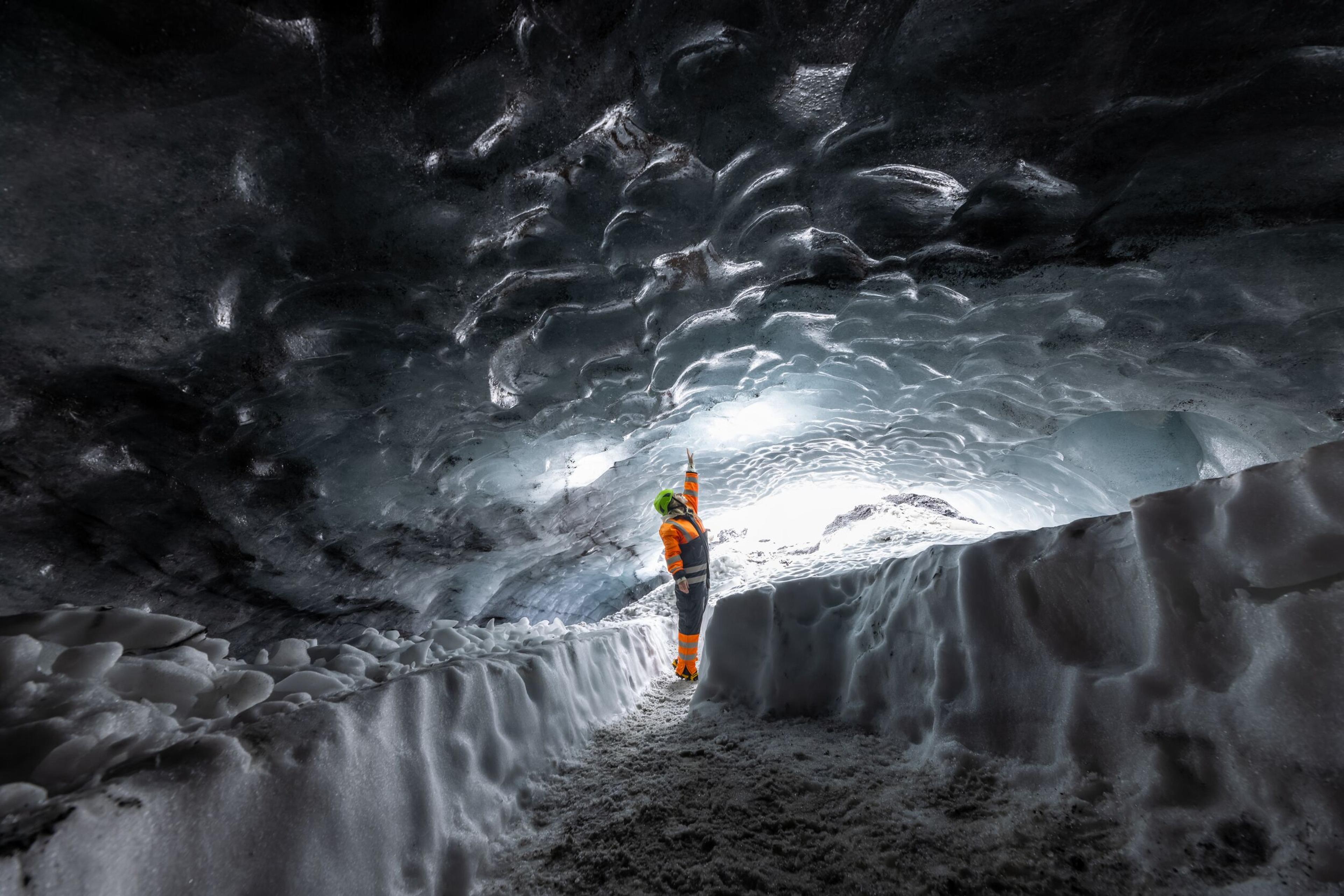 Person in orange and black suit exploring Askur ice cave, pointing at the illuminated ice ceiling, surrounded by snow.