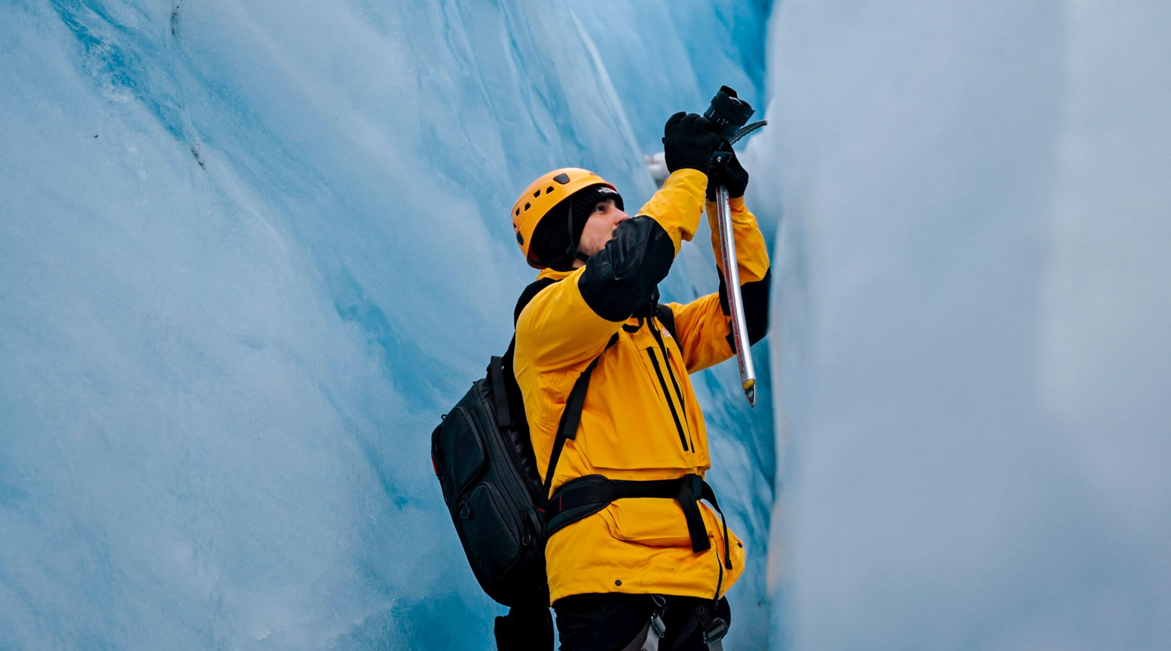 Person in yellow jacket and helmet holding an ice axe between blue ice walls.