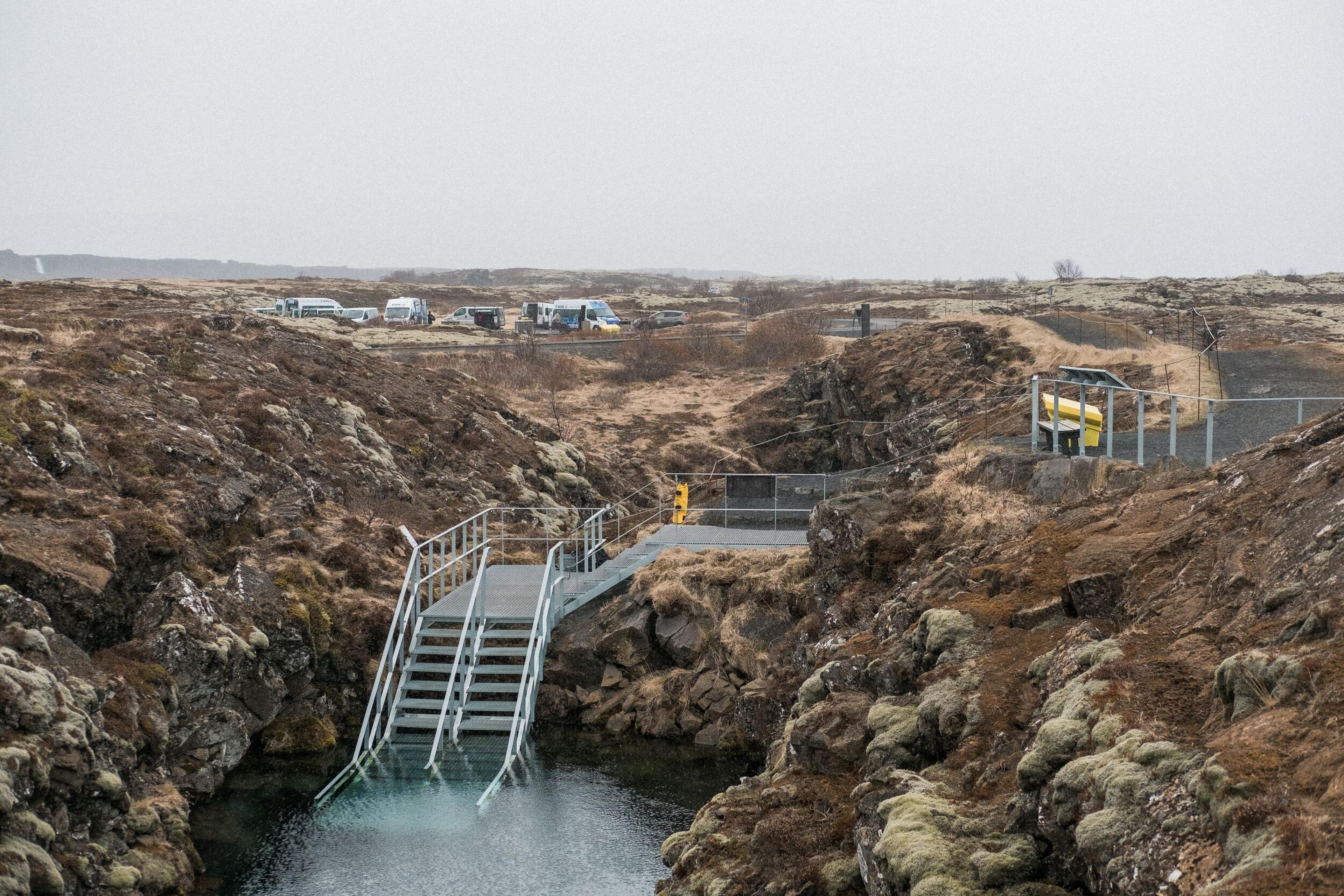 Metal stairs descend into clear water within a rocky fissure, surrounded by a mossy, barren landscape with campervans in the distance under an overcast sky.
