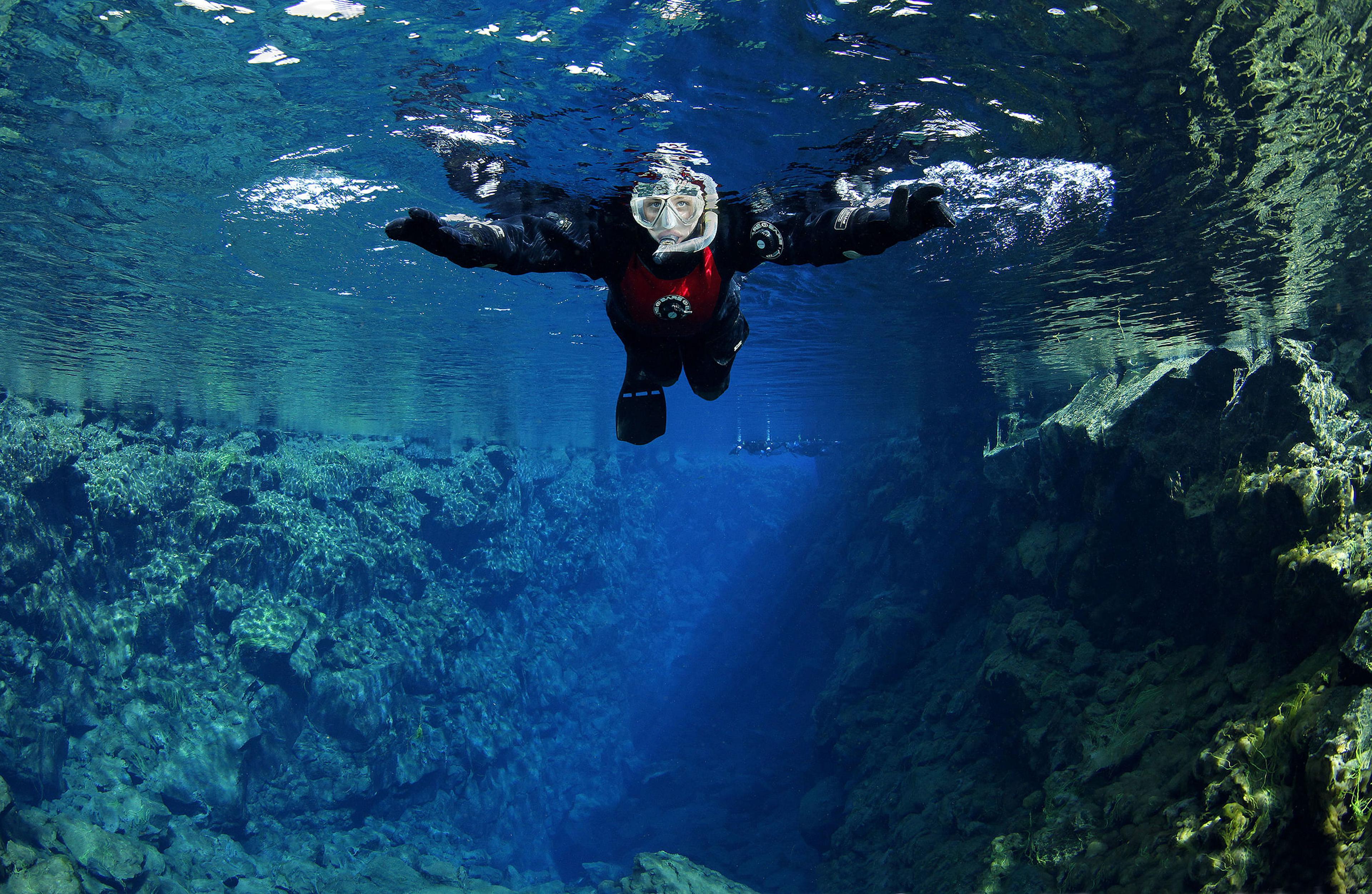 A snorkeler in a dry suit floats in crystal-clear blue water within an underwater rocky fissure.