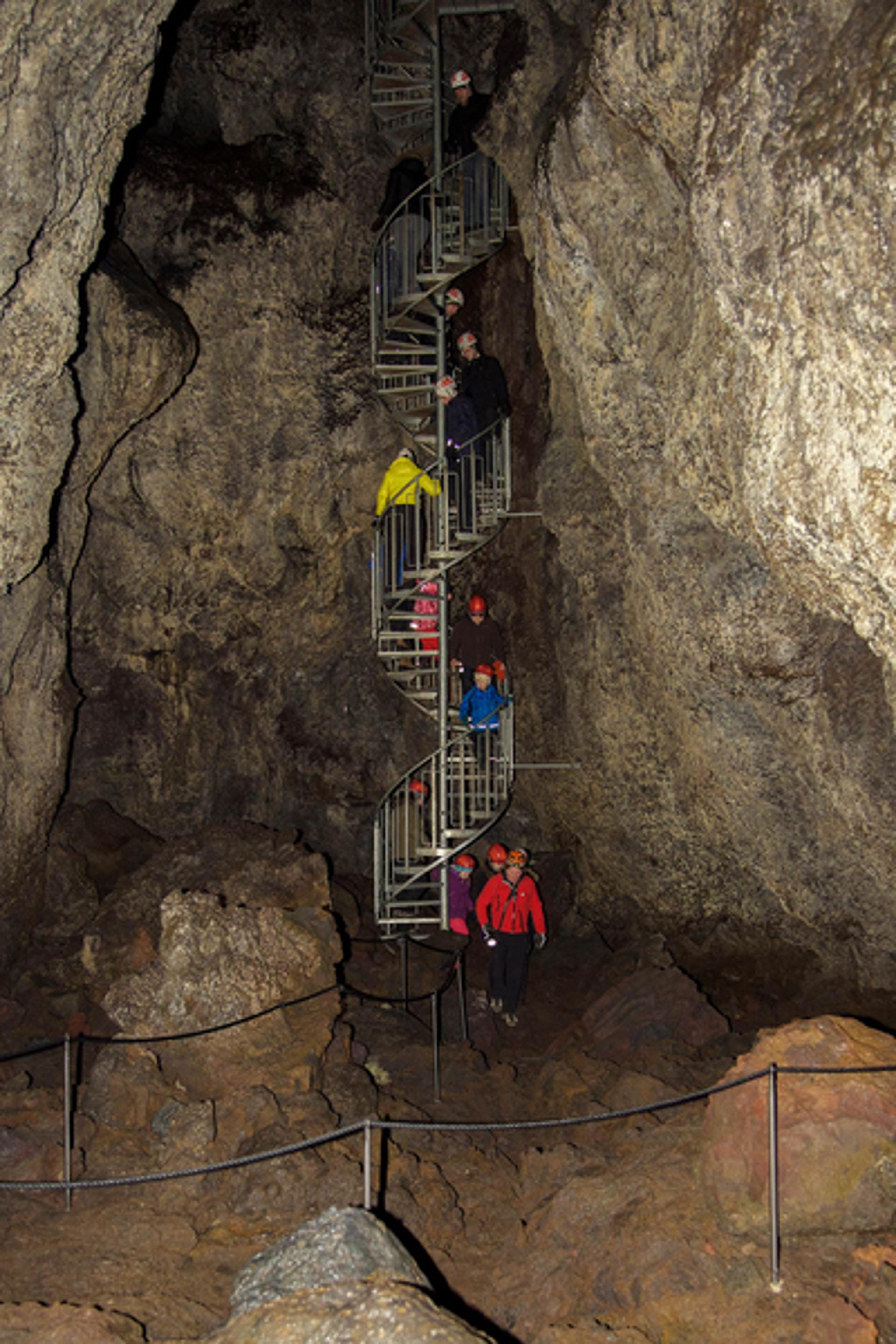 Tourists descend a steep metal staircase into the depths of a dark cave, exploring its rugged interior illuminated by sparse lighting.