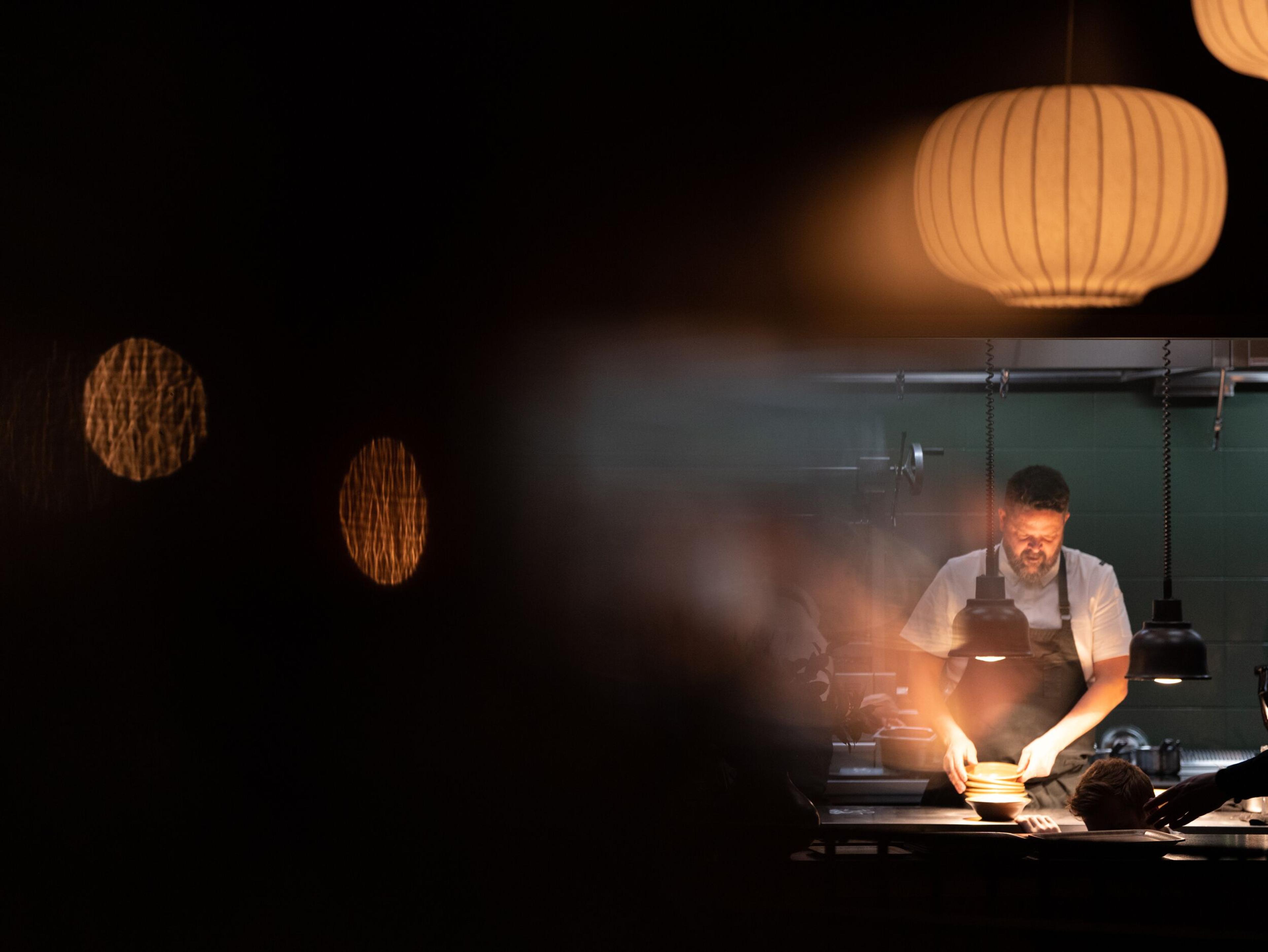 A chef works under bright task lights in a dark kitchen, with soft decorative lanterns hanging above.