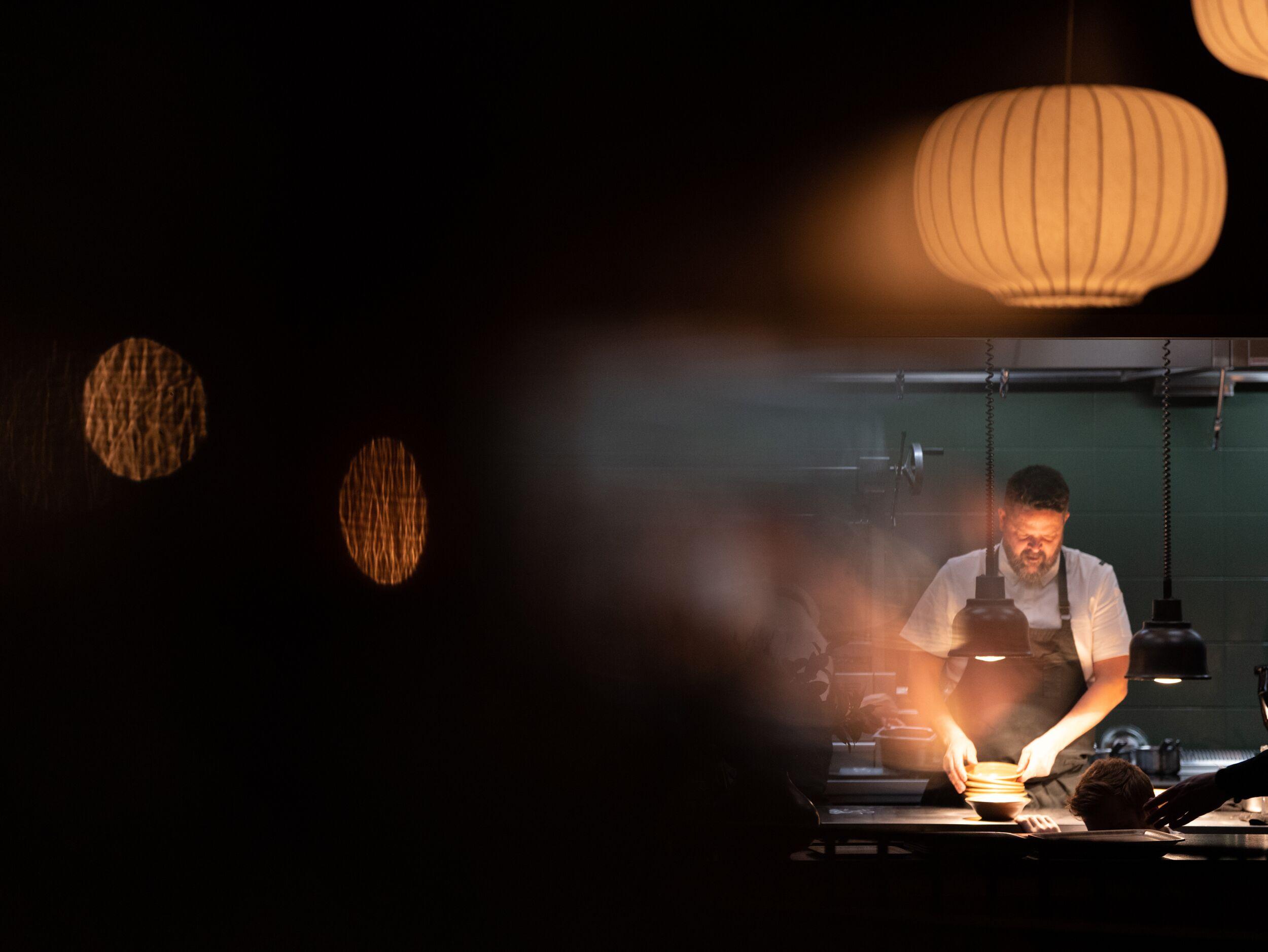 A chef works under bright task lights in a dark kitchen, with soft decorative lanterns hanging above.