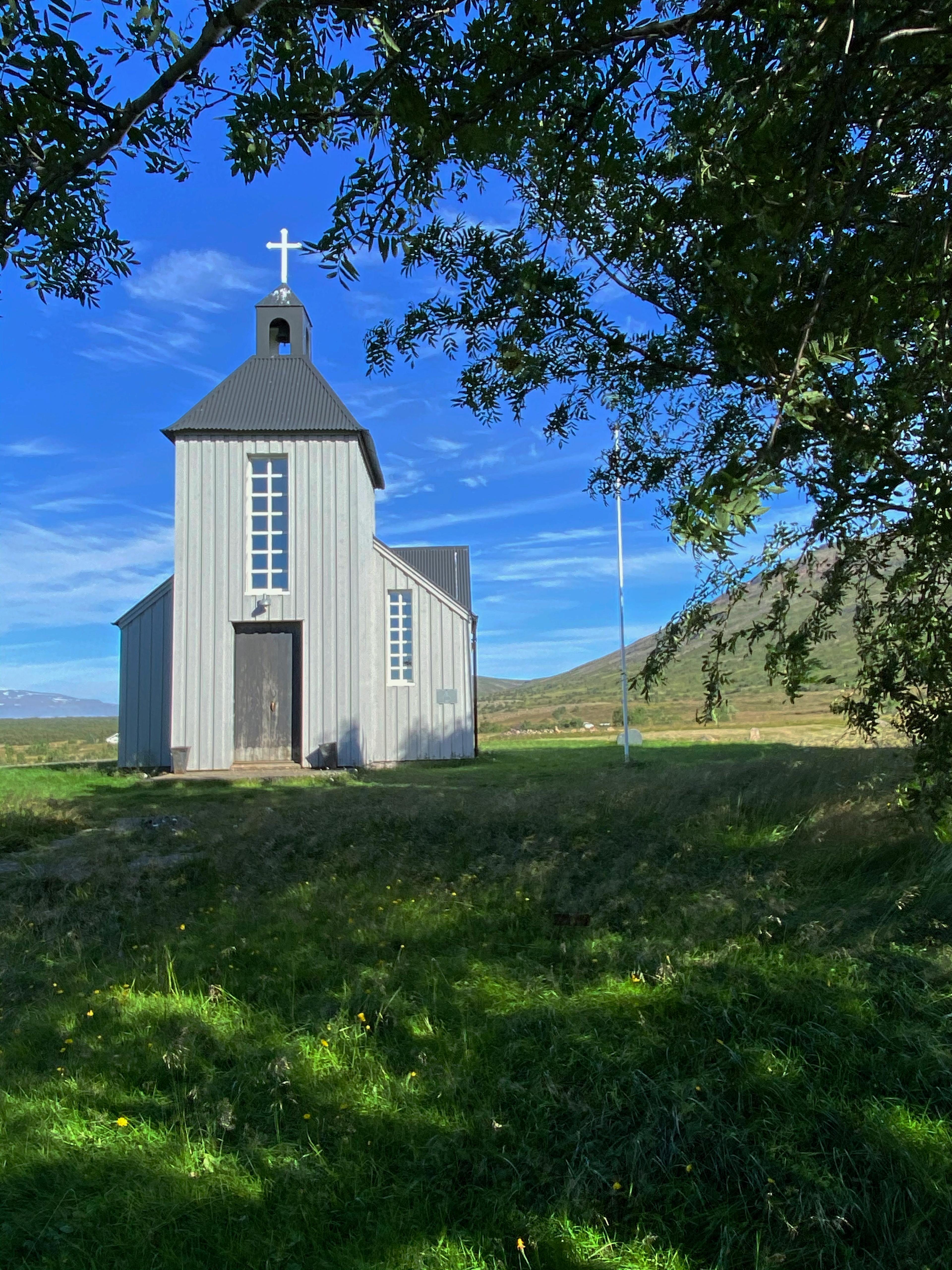 A charming small church in Húsafell, painted white with a black roof and cross, framed by green trees and a vibrant blue sky in a serene countryside setting.